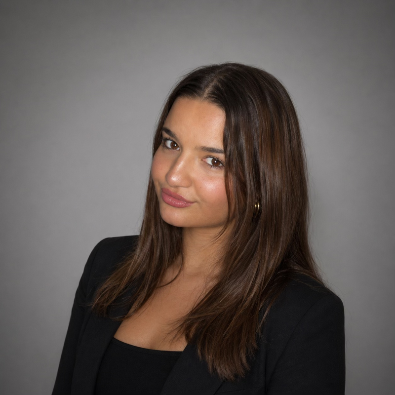 A young woman with long brown hair, wearing a black blazer and gold earrings, looking at the camera against a gray background.