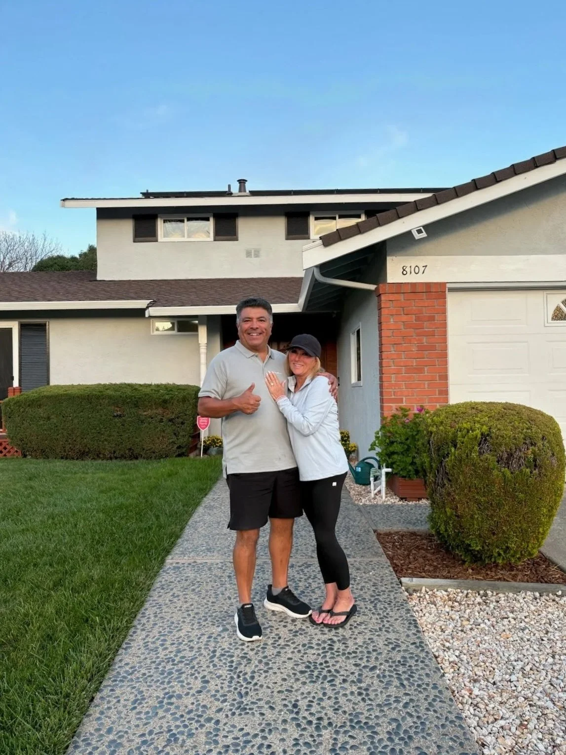 A smiling couple standing on a pebble walkway in front of a house, with the man giving a thumbs-up. The house has a white exterior, brick accents, and a two-story section in the background. The lawn is green and well-maintained.