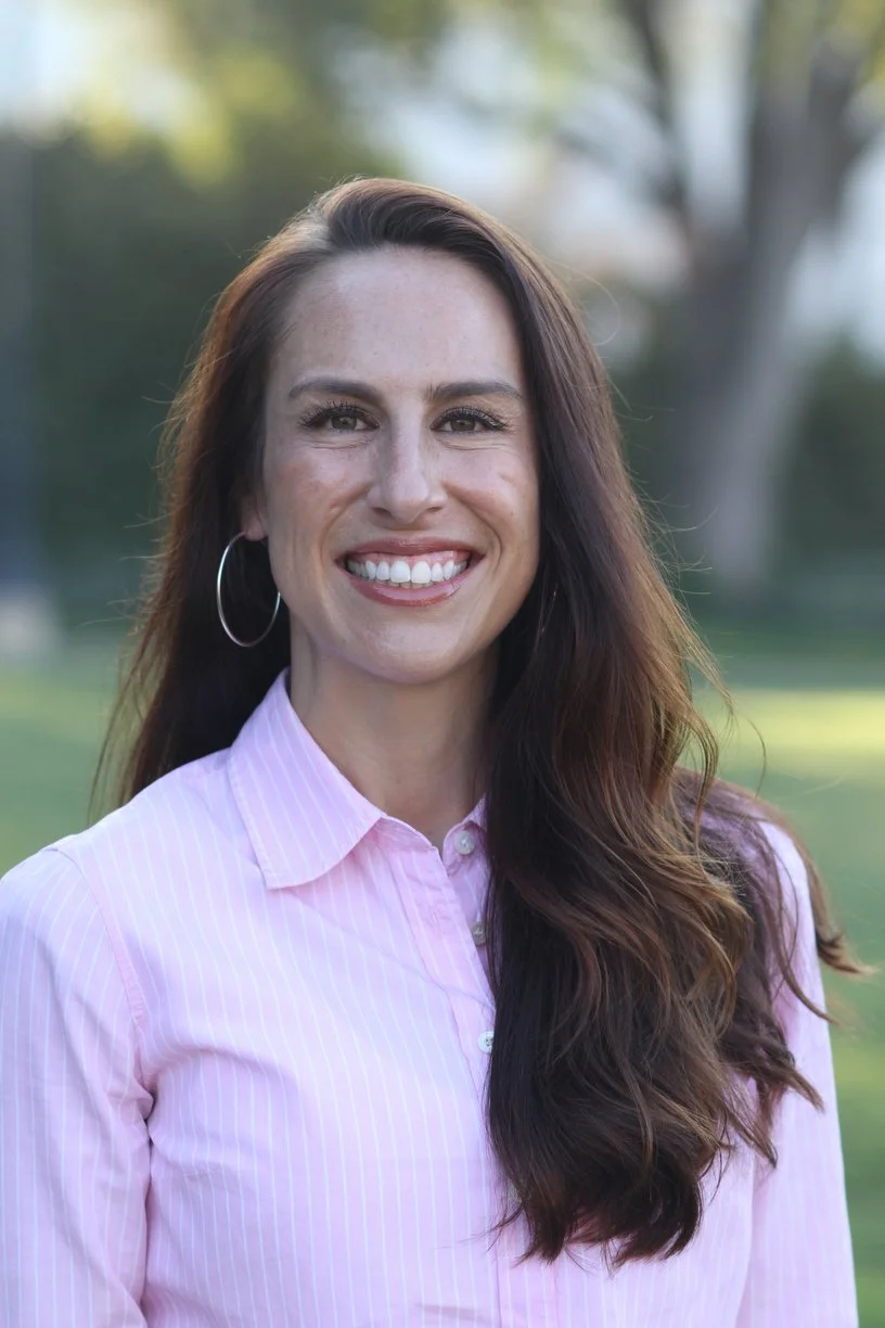 A woman with long brown hair, wearing hoop earrings and a pink striped button-up shirt, smiling outdoors with blurred trees in the background.