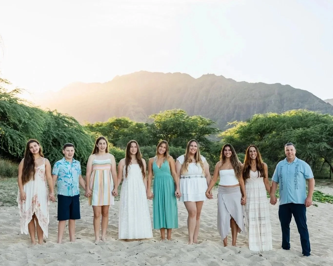 Family holding hands on a beach during sunset, with trees and mountains in the background.