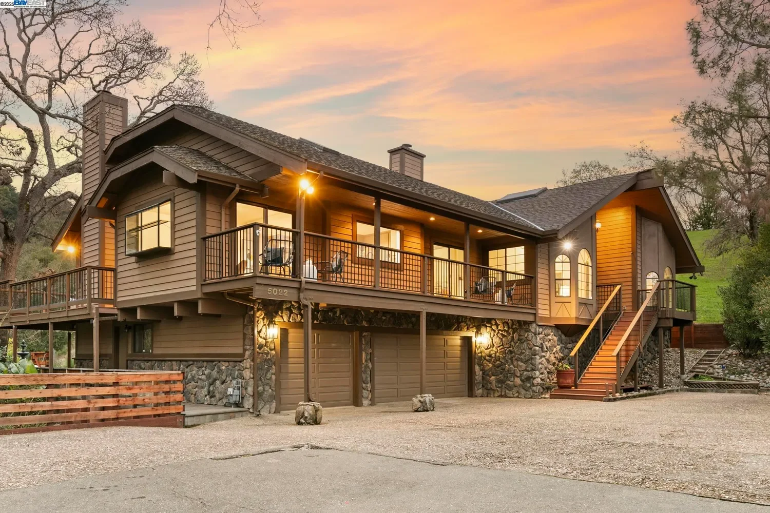 A two-story house with a front driveway and three garage doors underneath a large balcony with outdoor furniture, surrounded by trees and hills at sunset.