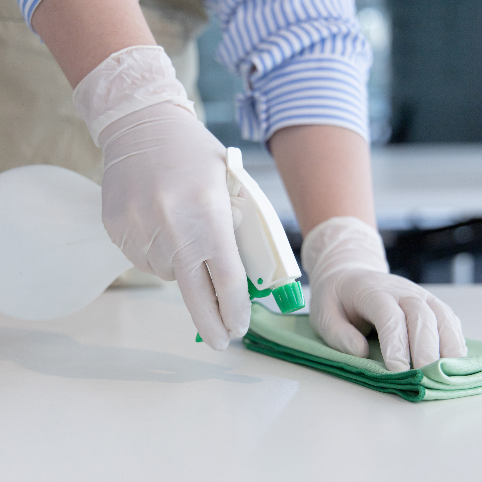Person disinfecting a countertop with a spray bottle and microfiber cloth while wearing protective gloves.