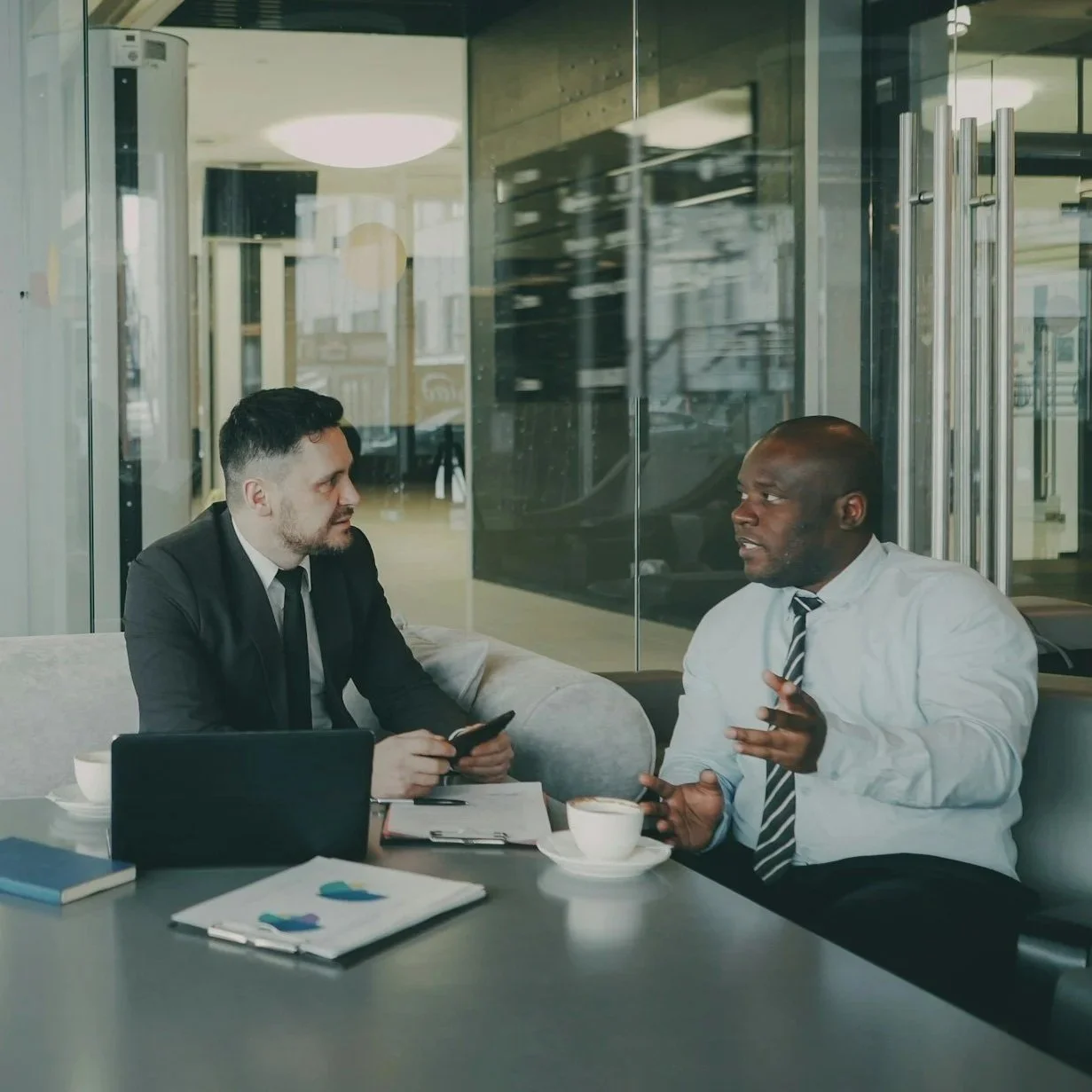 Two business professionals having a discussion in a modern office lounge, with laptops, documents, cups of coffee, and one person holding a smartphone.
