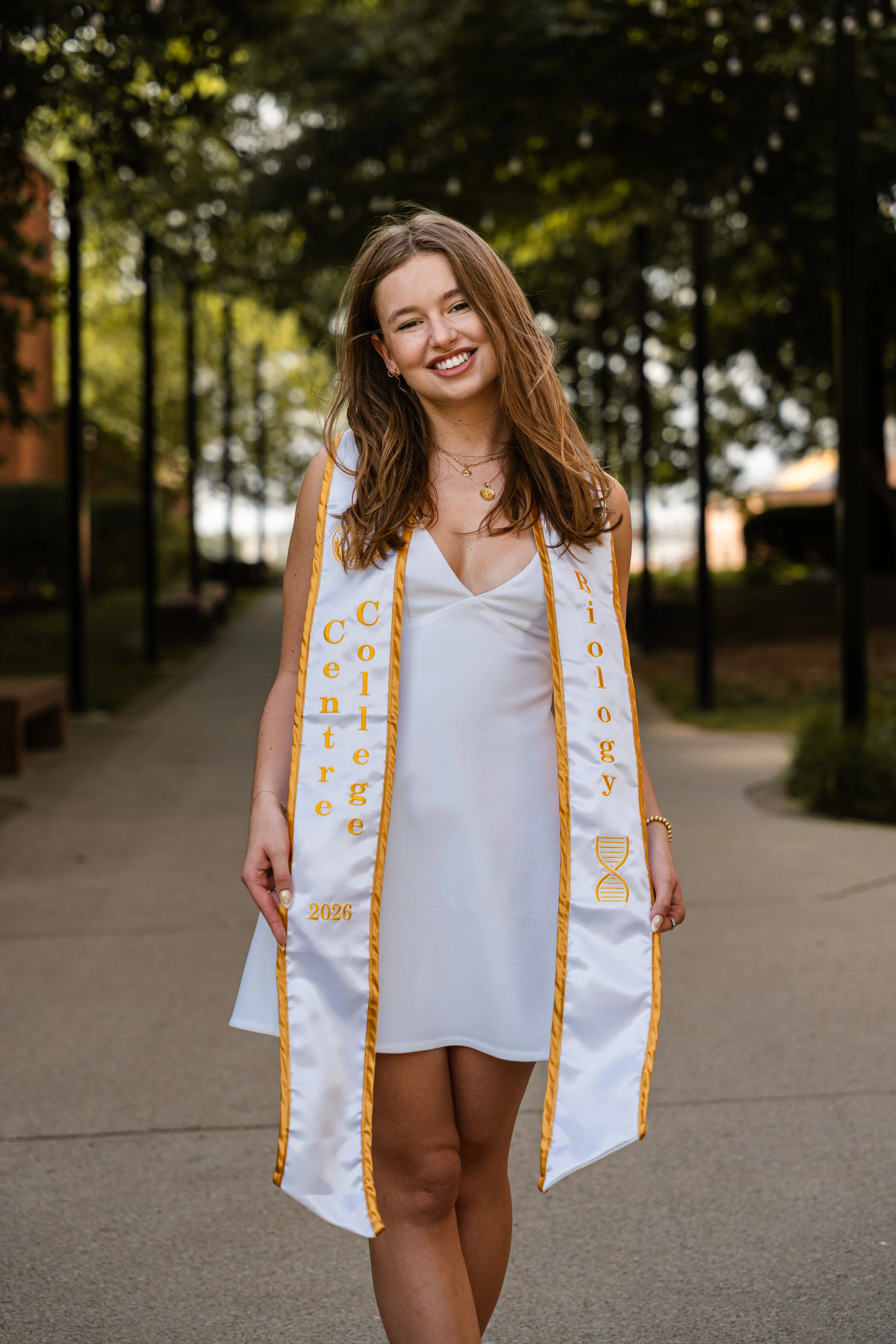 A young woman in a white dress and graduation stole standing outdoors on a pathway, smiling and looking at the camera. The stole has gold trim and text, and the year 2026 is embroidered on it.