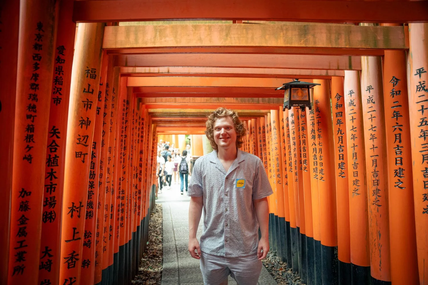 A smiling man standing in a pathway lined with orange torii gates with black inscriptions, in a Japanese shrine or temple.