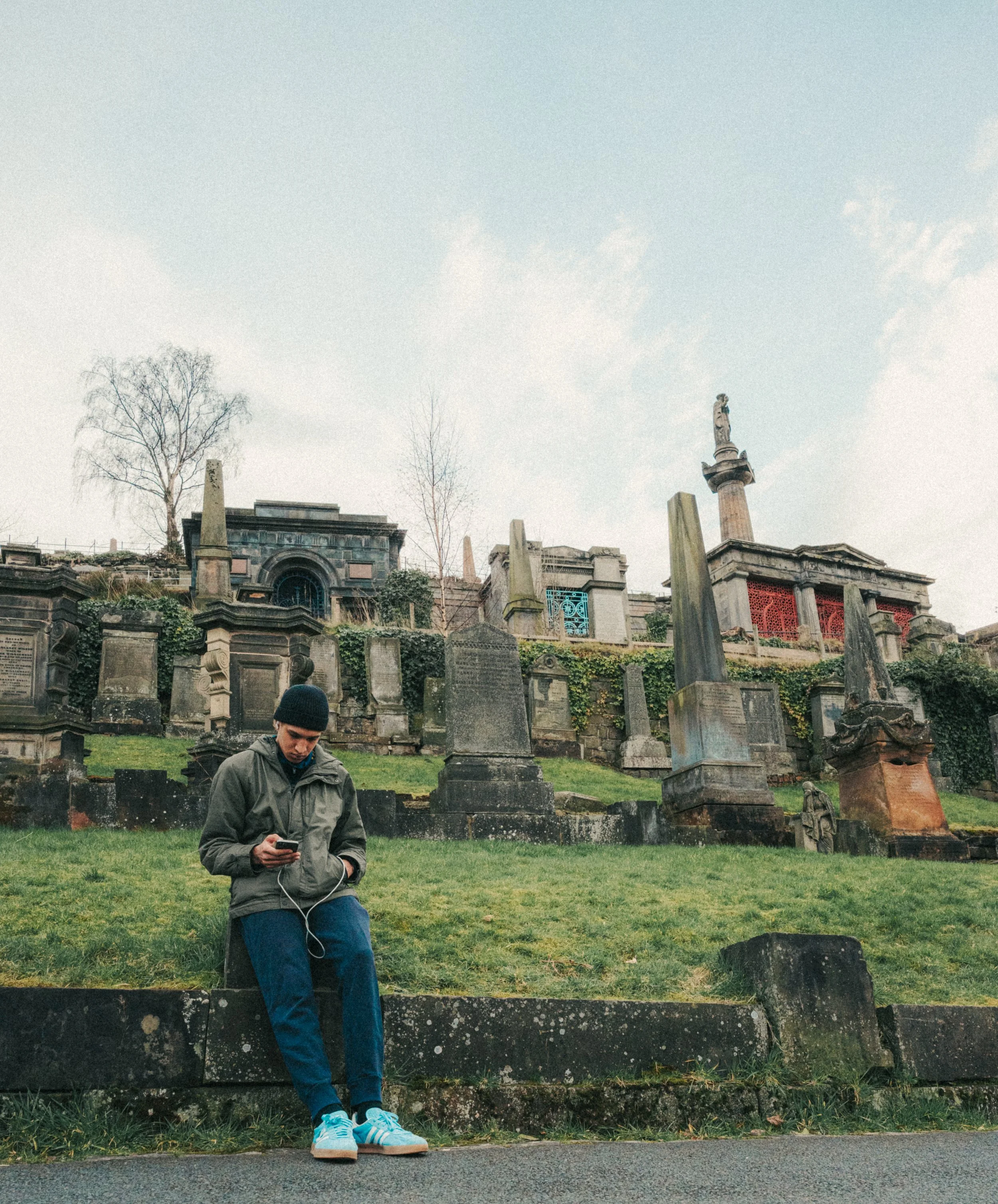 A young man in a gray jacket and blue pants leans against a low wall, looking at his phone, in front of a cemetery with tombstones and statues on a cloudy day.
