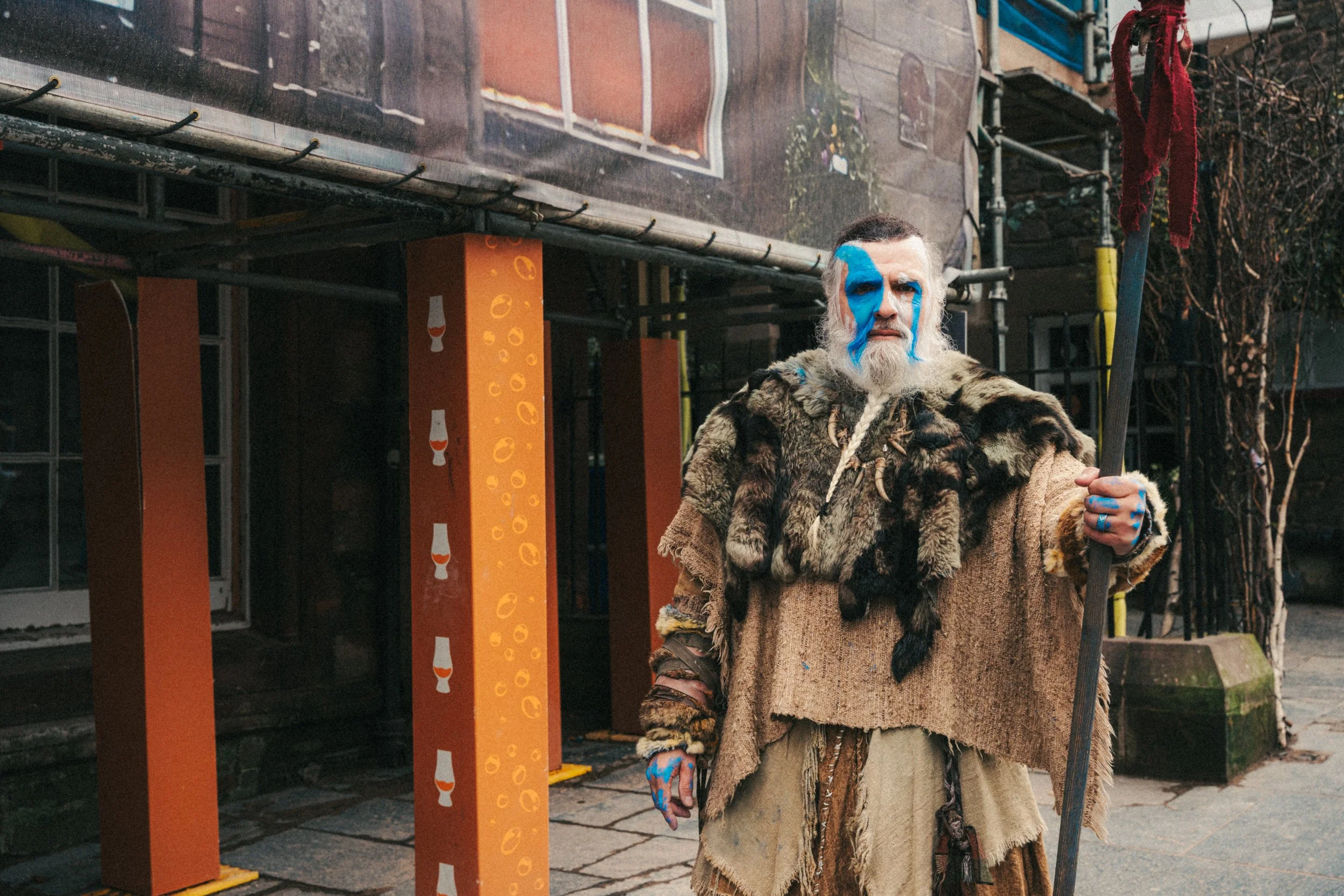 A man in tribal costume with blue facial paint and fur clothing, holding a staff, standing outside on a city street.