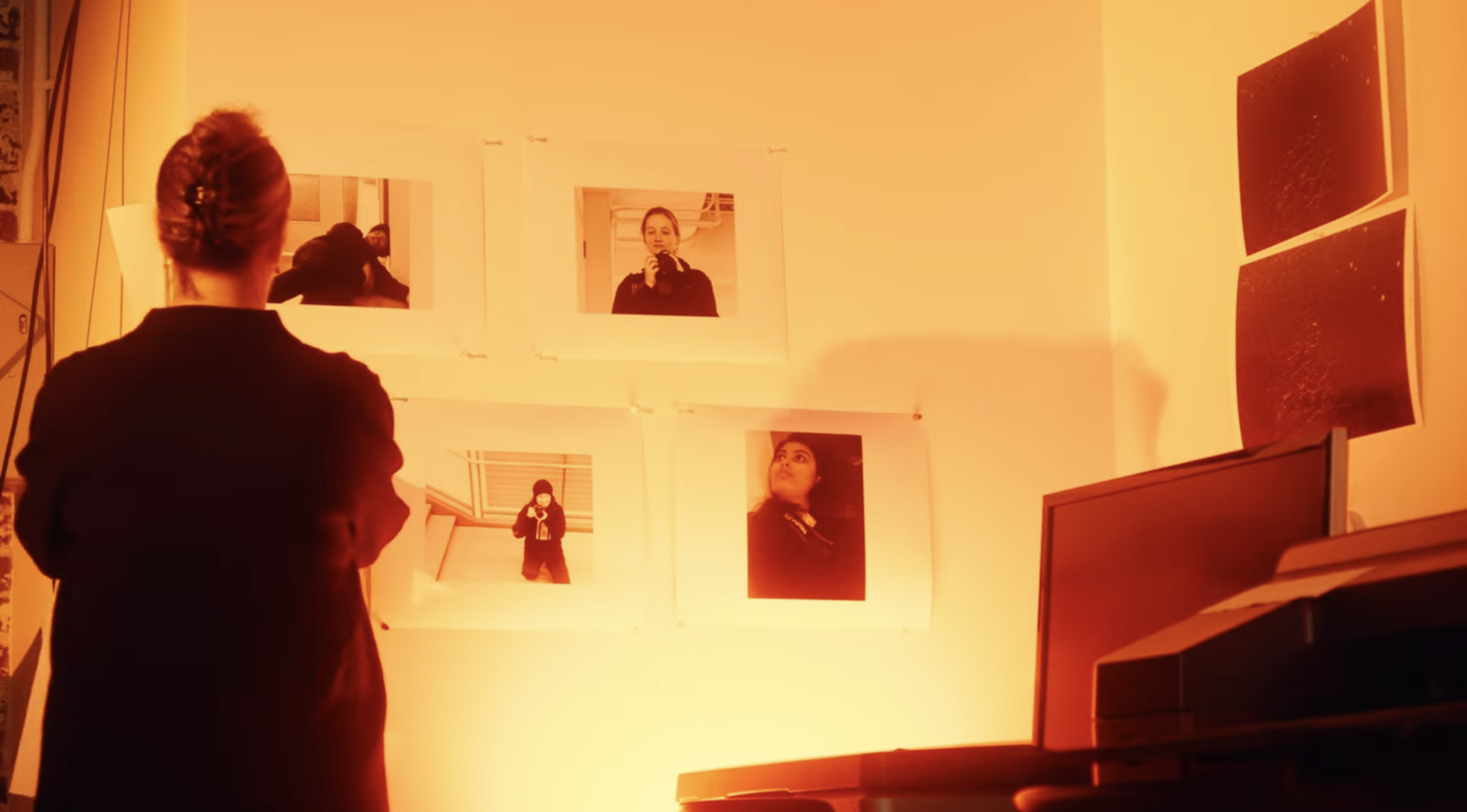 A woman viewed from behind, sitting and looking at a wall decorated with multiple framed photographs, with orange ambient lighting.