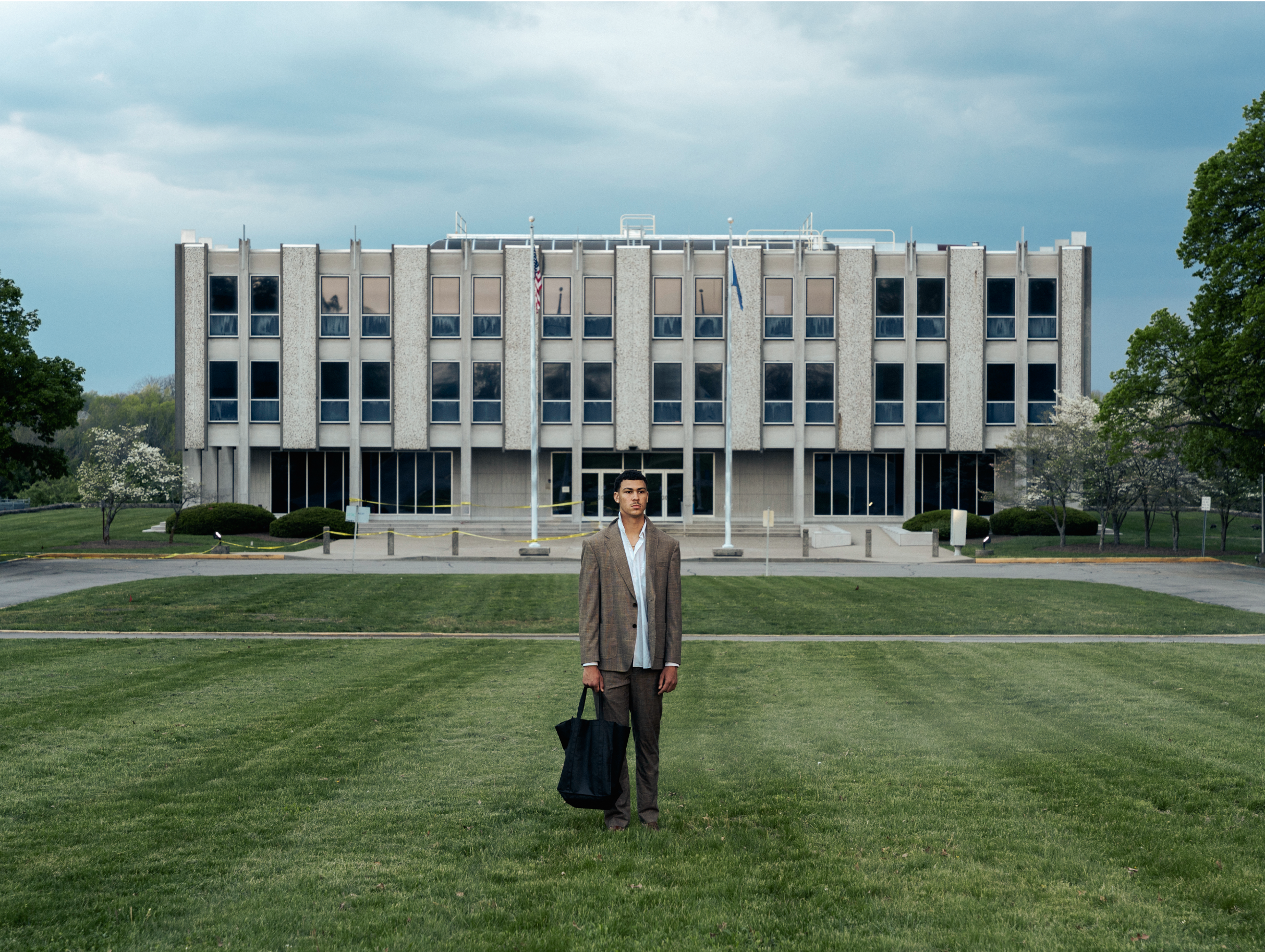 A man in a brown suit holding a black bag walks across a grassy area in front of a large office building. The building has multiple windows and flagpoles with flags in front of it. The sky is cloudy.