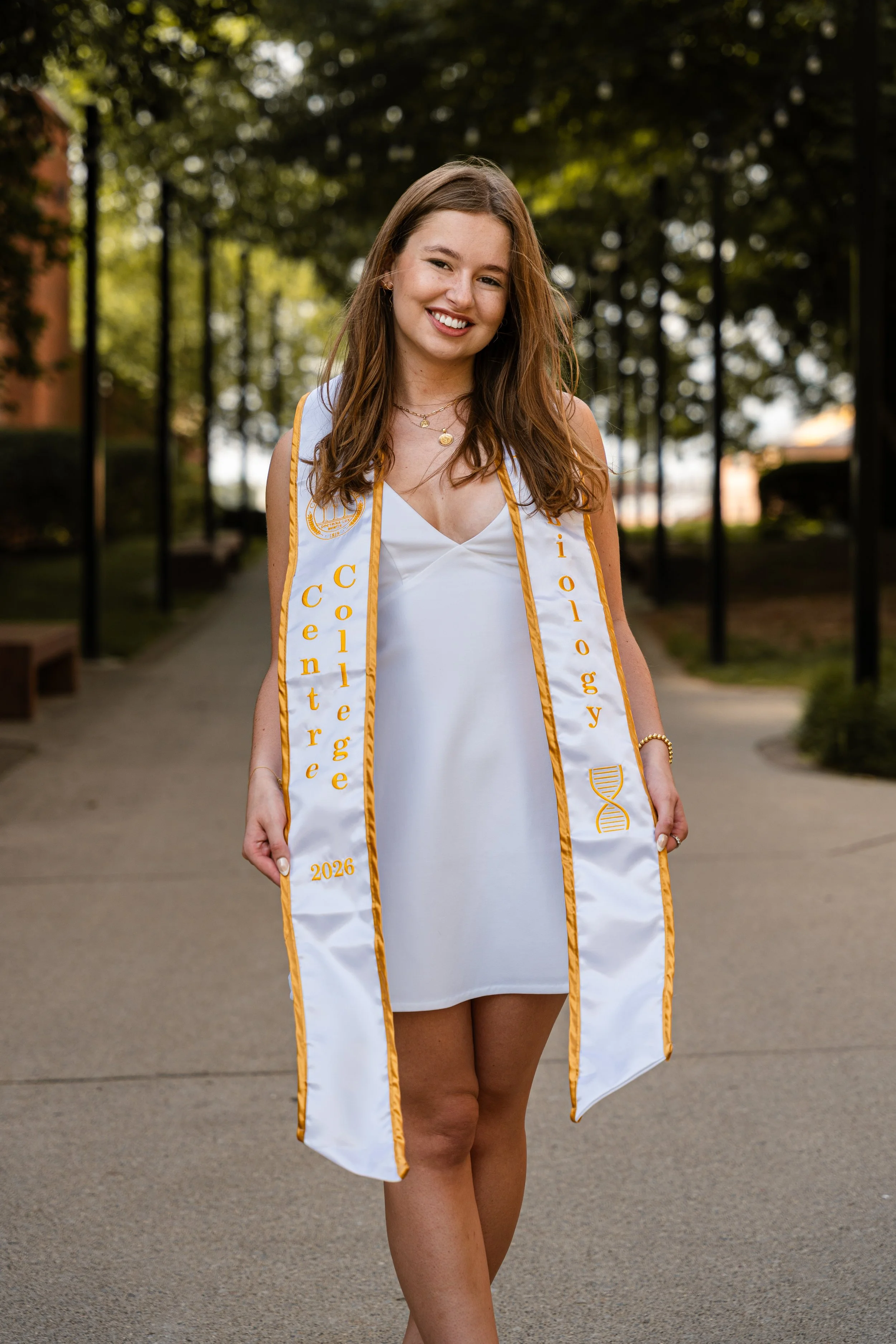 A young woman in a white dress wearing a white and gold graduation stole, smiling outdoors on a pathway lined with trees.