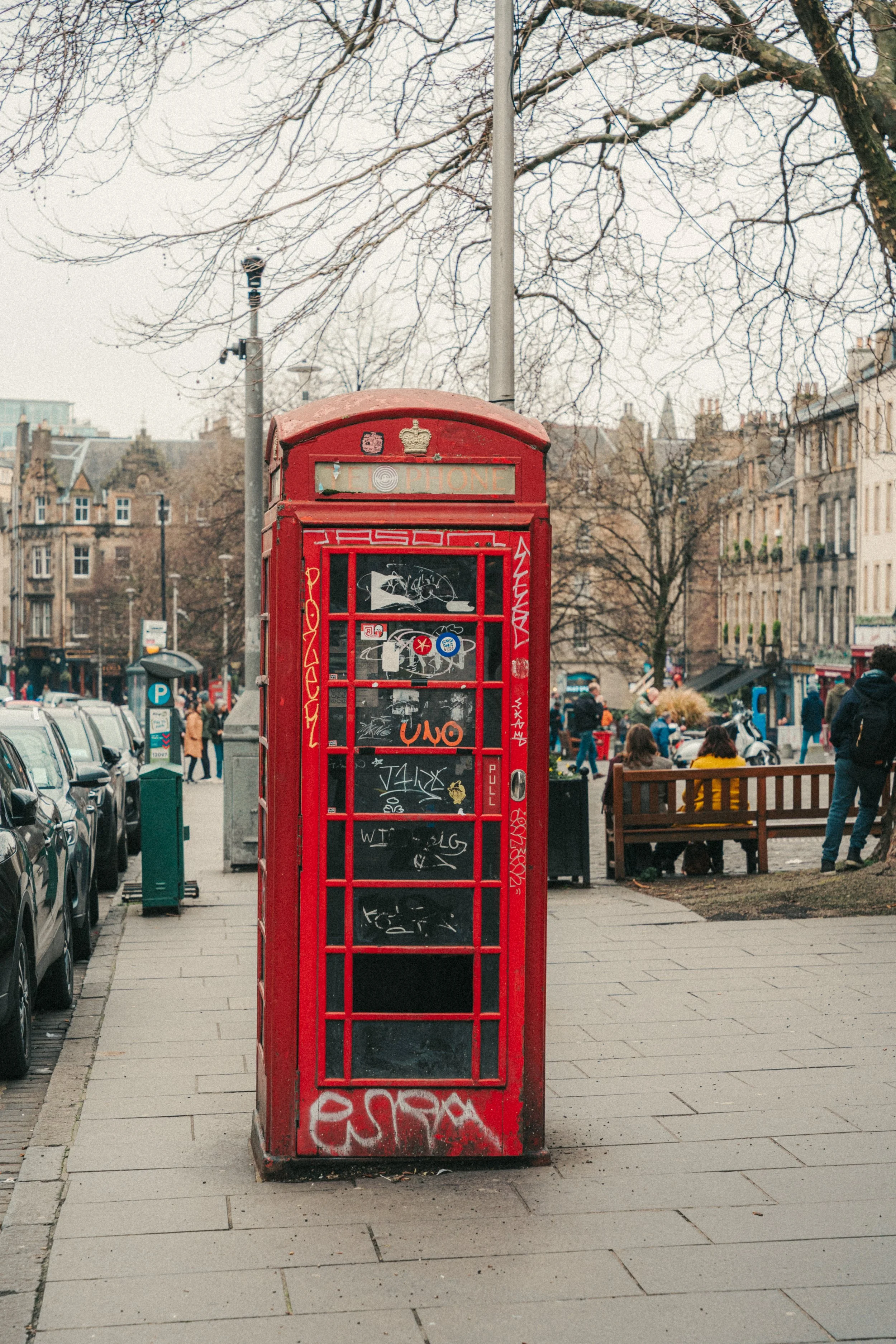 Red British telephone booth covered in graffiti, with a city street scene in the background, including parked cars, pedestrians, and buildings with trees lining the street.