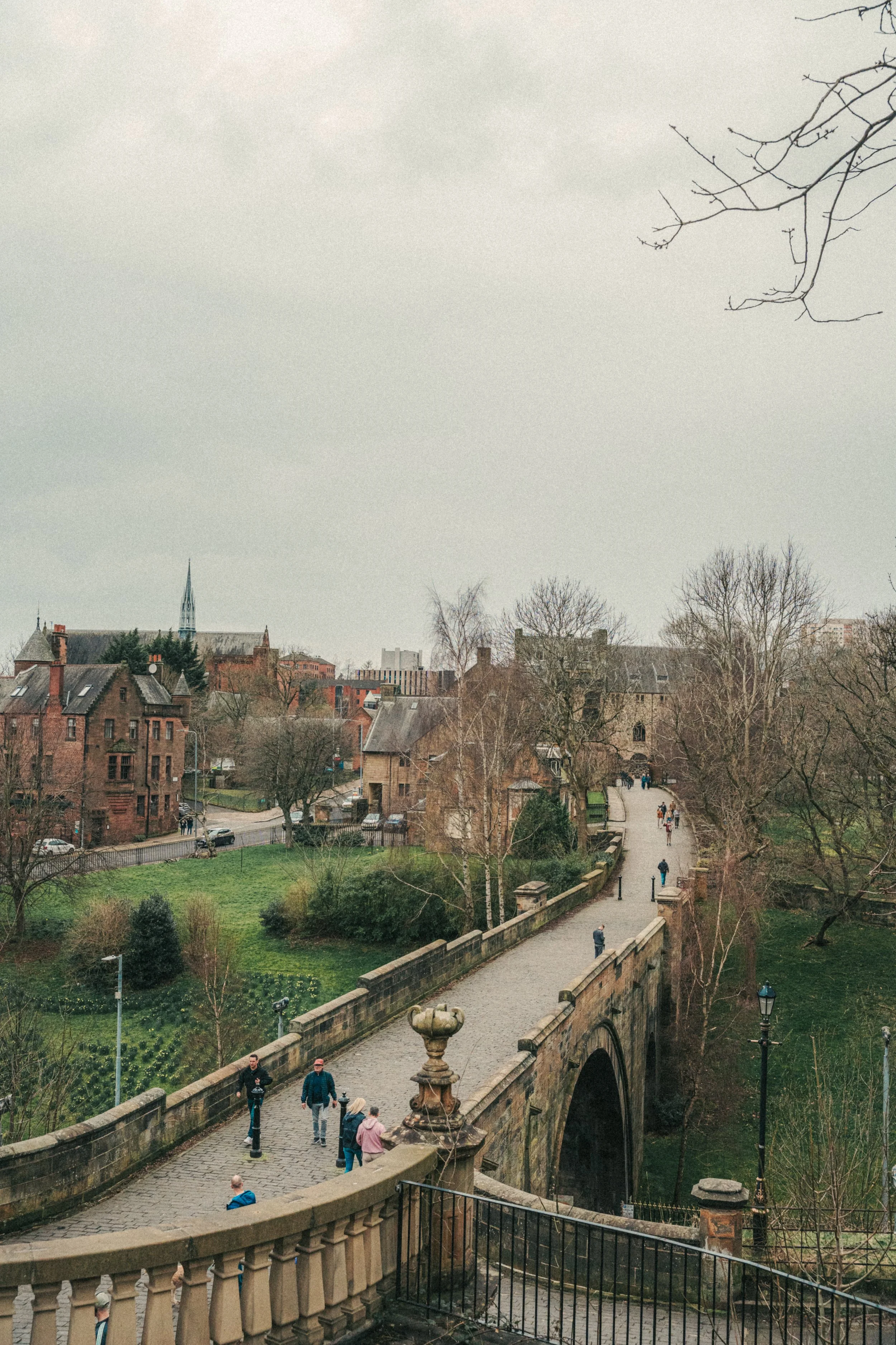 A stone bridge with a balustrade over a small river, with people walking on it, leading to a park with grass, trees, and buildings in the background. The sky is overcast.