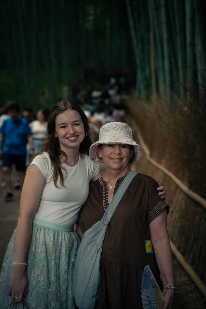 Two women smiling and standing together in a bamboo forest, with many people in the background walking along a trail.