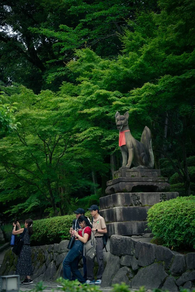 A large stone statue of a fox with a red bib sits on a stone pedestal in a lush green park. Three people are observing and taking photos of the statue, surrounded by dense trees and bushes.