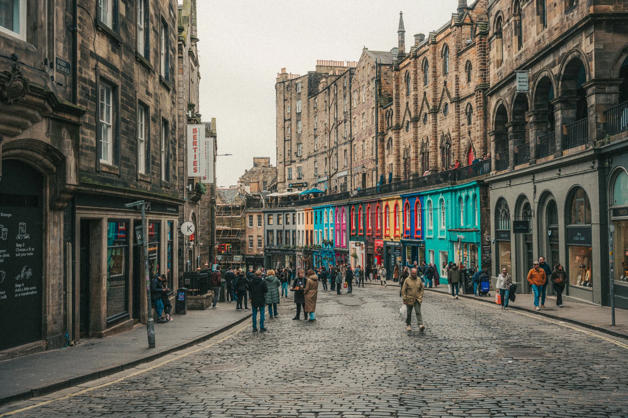 A crowded cobblestone city street with older stone buildings, cafes, and colorful storefronts, with people walking and socializing outdoors.