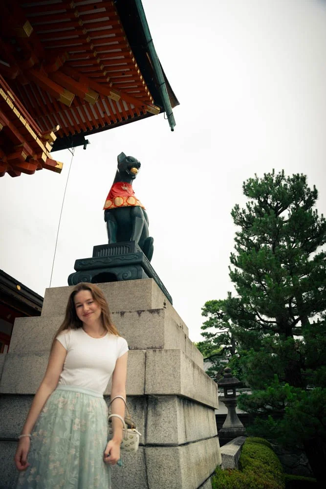 Young woman standing in front of a stone pedestal with a black dog statue wearing a red collar, part of a traditional Japanese temple or shrine, with green trees and a pagoda roof in the background.