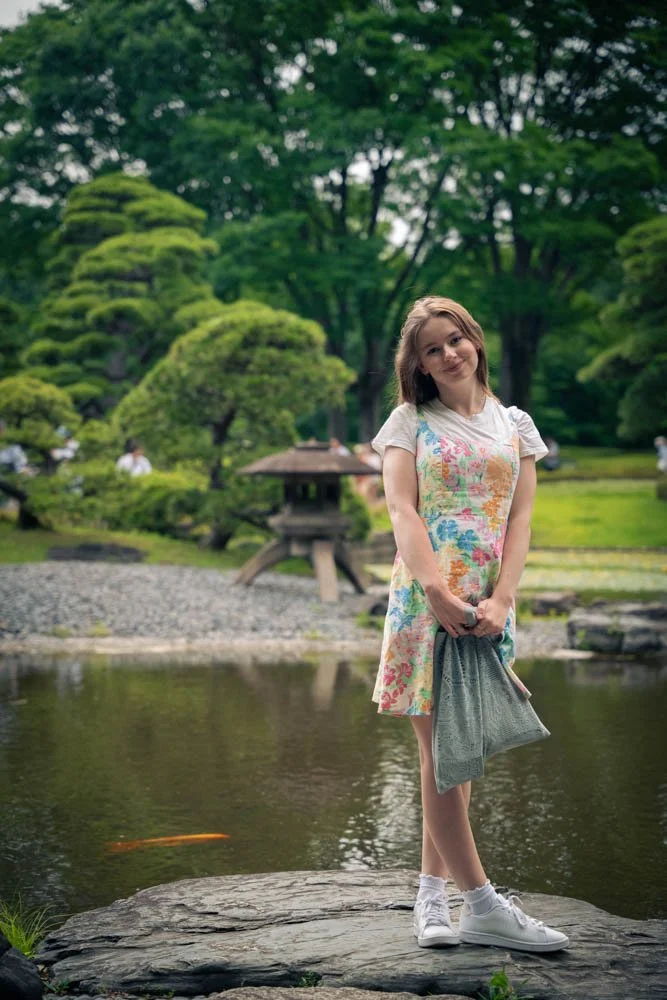 A young woman in a floral dress and white sneakers standing on a rock near a pond in a lush garden.