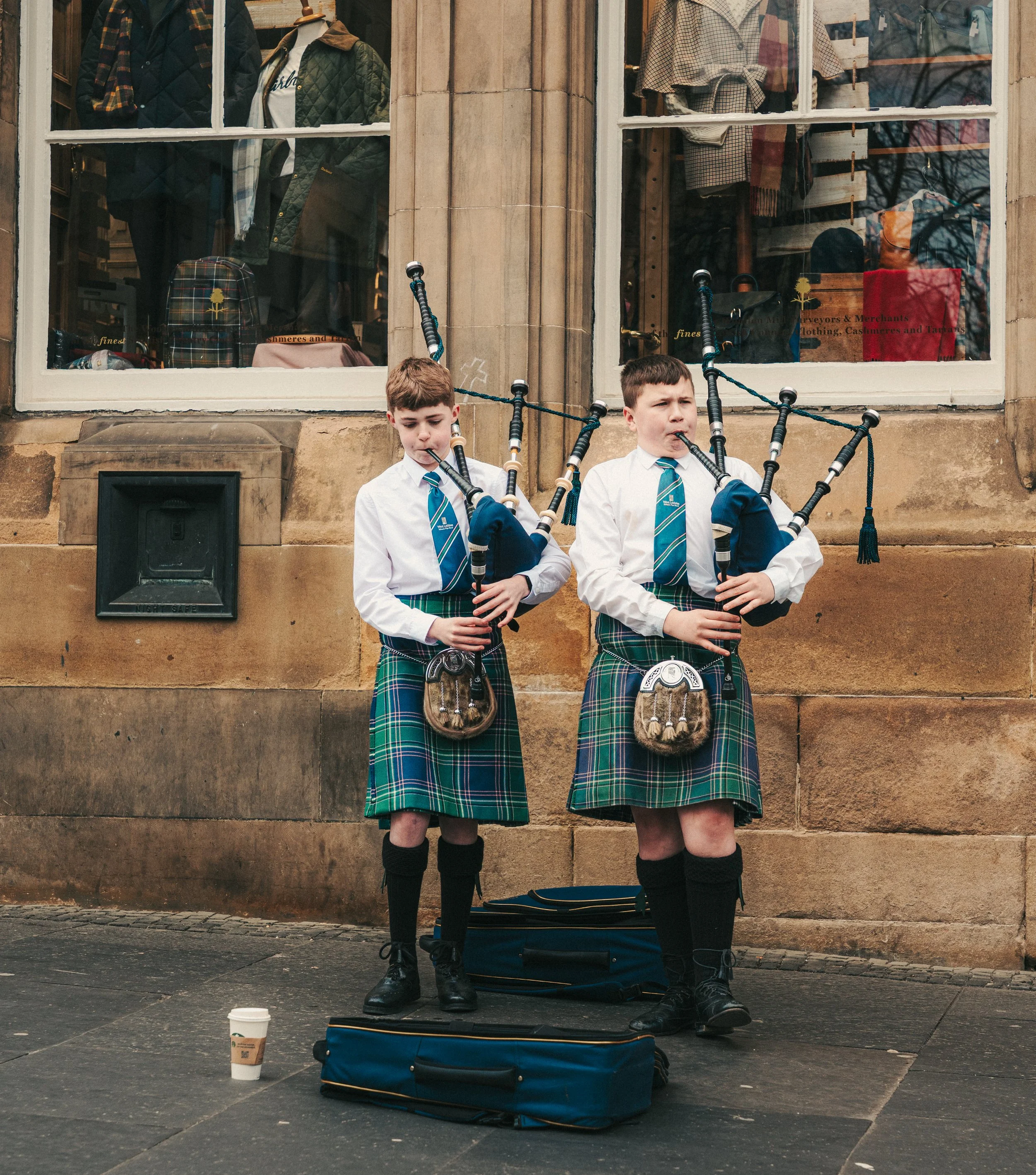Two young boys playing bagpipes in front of a school or public building, wearing school uniforms with tartan skirts and striped ties, bagpipe bags on the ground, and a coffee cup nearby.