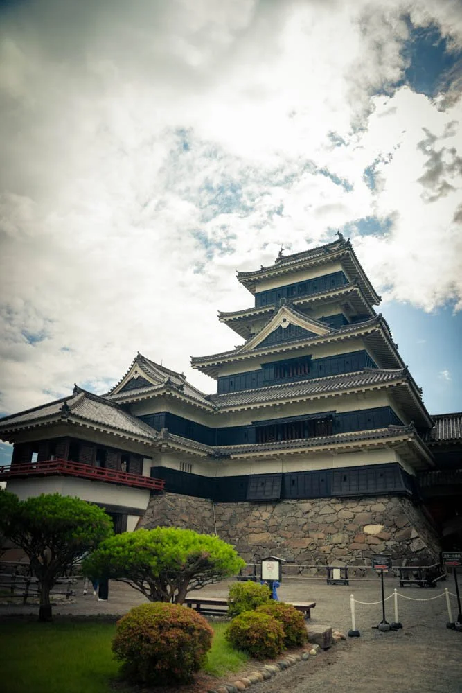 A traditional Japanese castle with multiple tiers, black and white exterior, and a stone foundation, under a cloudy sky.