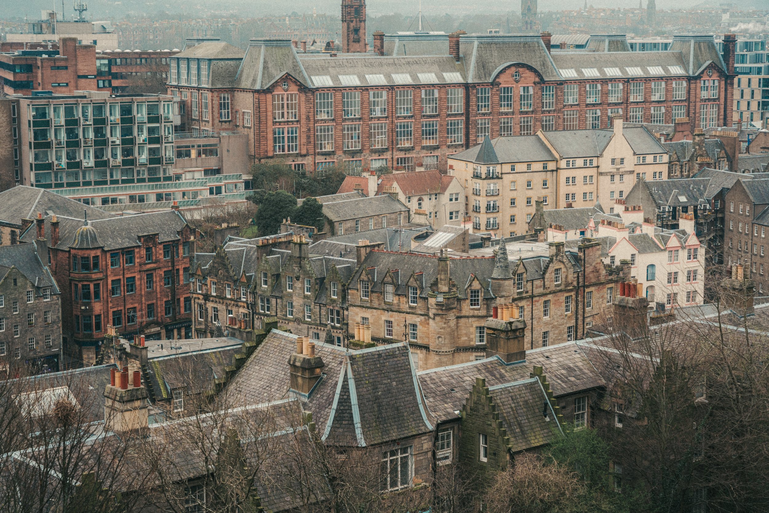 A cityscape featuring a mix of historic and modern buildings with rooftops, windows, and chimneys visible, under a hazy sky.