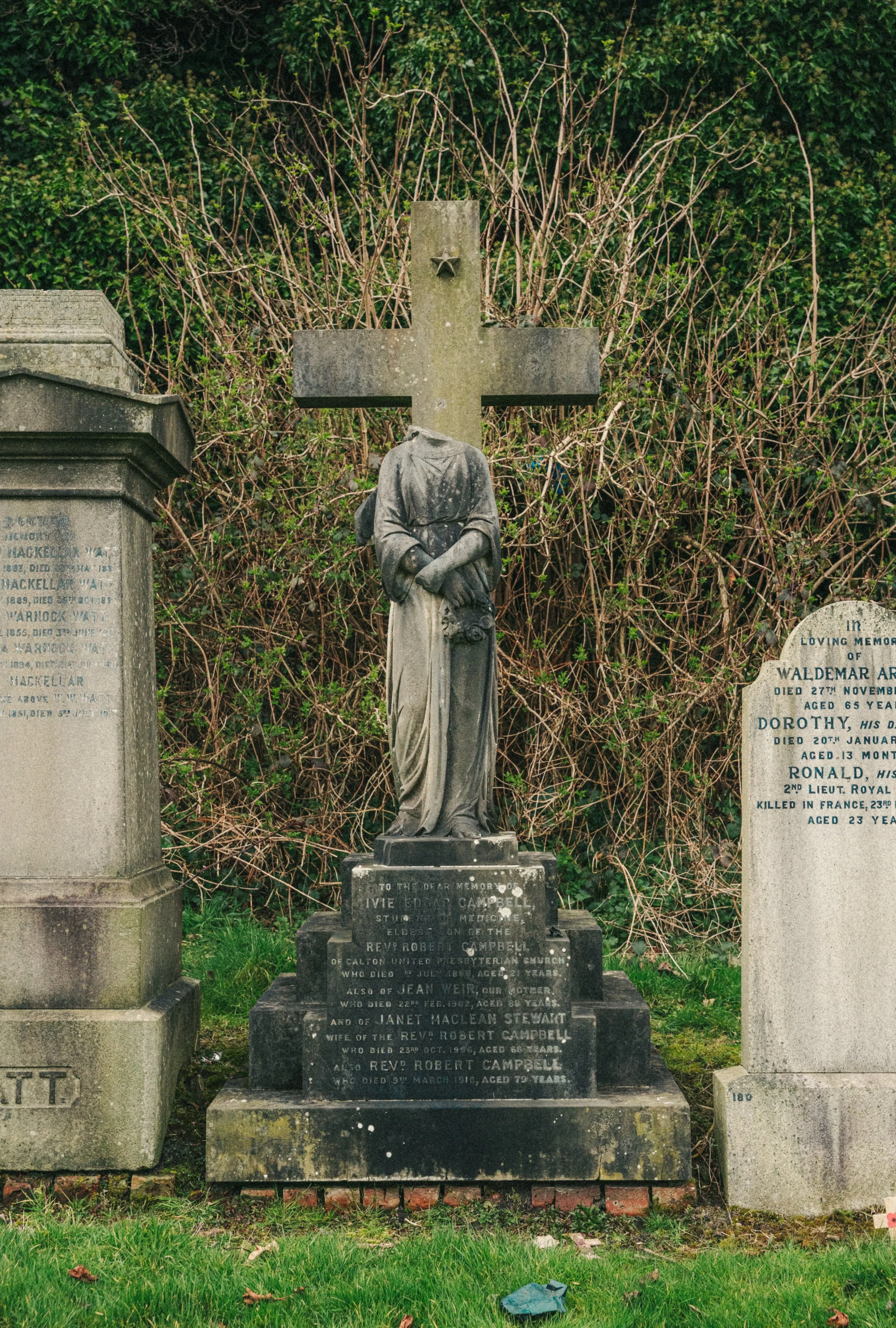 Gravestone with a cross-shaped top and a statue of a woman with head bowed and hands folded, situated between two other gravestones, in a graveyard with greenery and bushes in the background.