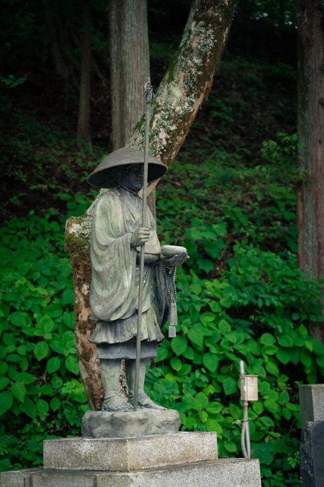 A weathered stone statue of a samurai warrior dressed in traditional clothing, holding a sword and a bowl, standing in a lush green forest. The statue has a wide-brimmed hat and is mounted on a stone pedestal.