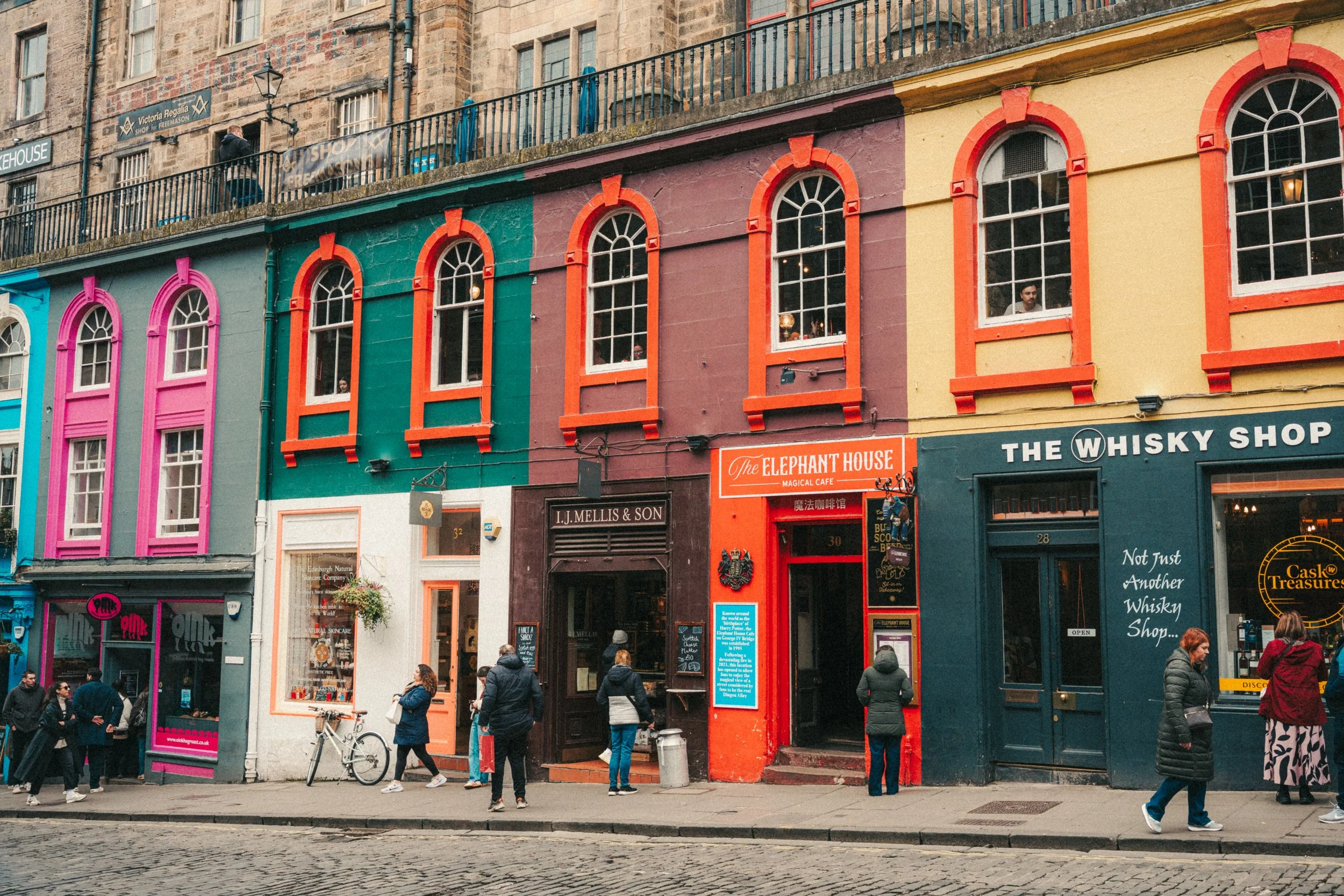 Colorful building facades in a city street with pedestrians walking and standing, shops and signs visible, overcast weather.