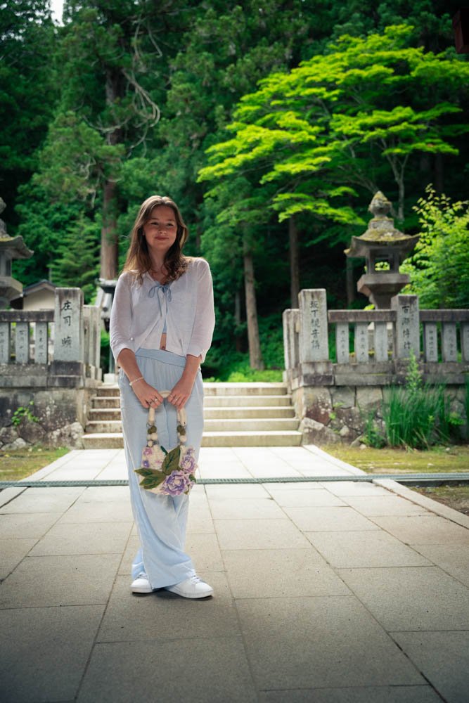 A woman standing on an outdoor stone walkway in front of a stone staircase and traditional Japanese lanterns, surrounded by lush green trees.