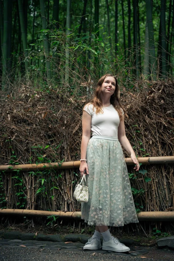 A young woman with long hair standing in front of a bamboo fence and lush greenery in a forest. She is wearing a white t-shirt, a light-colored tulle skirt with floral embroidery, white sneakers, and holding a small purse.