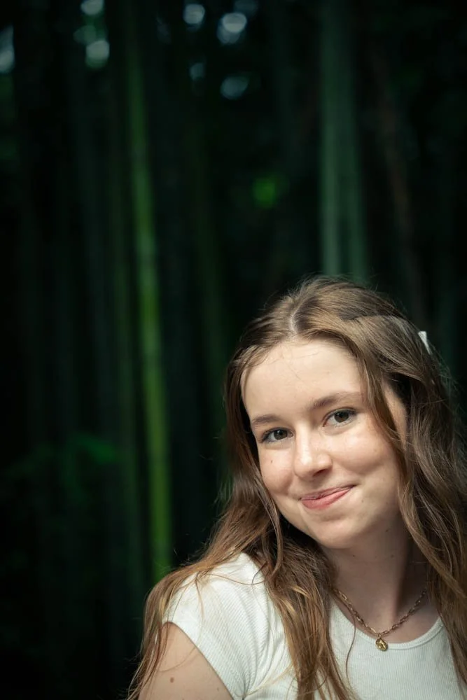 A young woman with long, wavy brown hair smiling and sticking out her tongue, standing in front of a green bamboo forest.