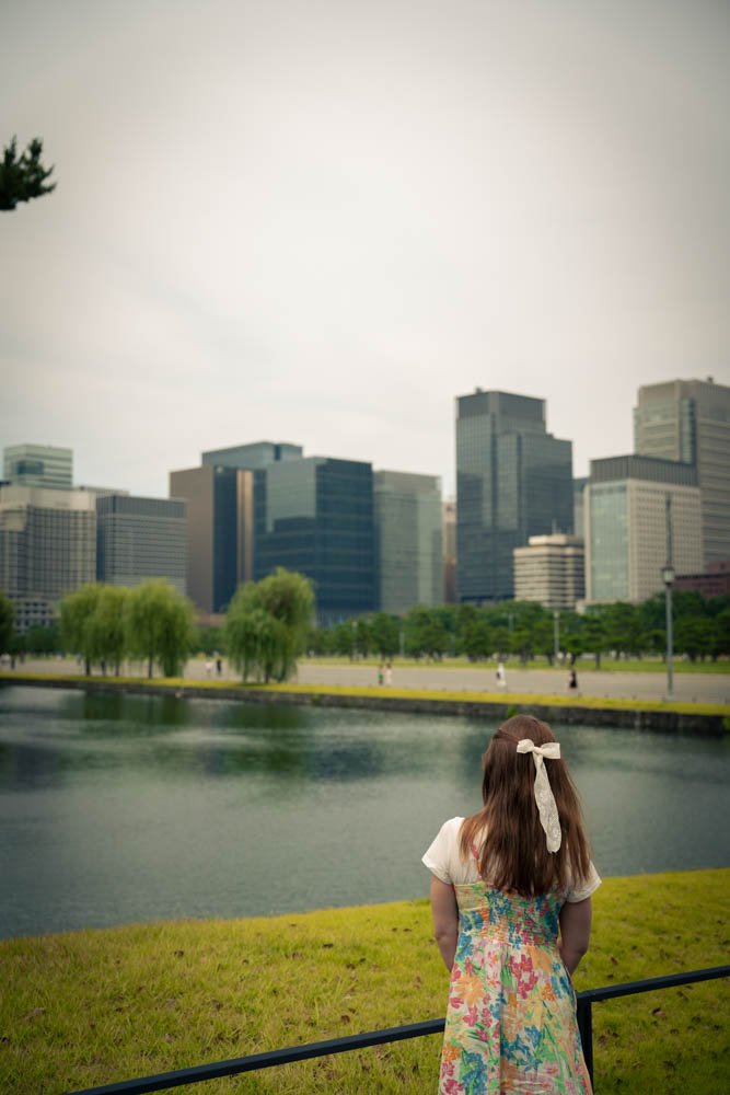 A young girl with brown hair and a white bow, wearing a colorful floral dress, stands by a lake looking at a cityscape with modern skyscrapers and green trees in the background.