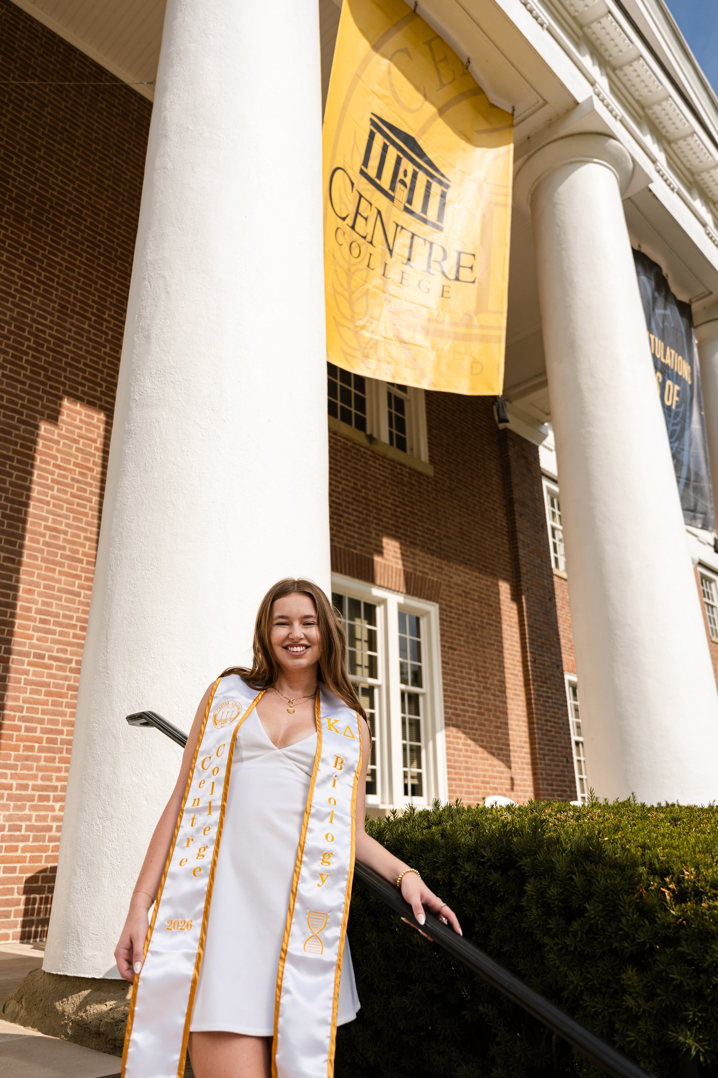 A young woman in a white dress and a graduation stole standing in front of a university building with large white columns and a banner that reads 'Centre College.' She is smiling and holding onto a railing with one hand.