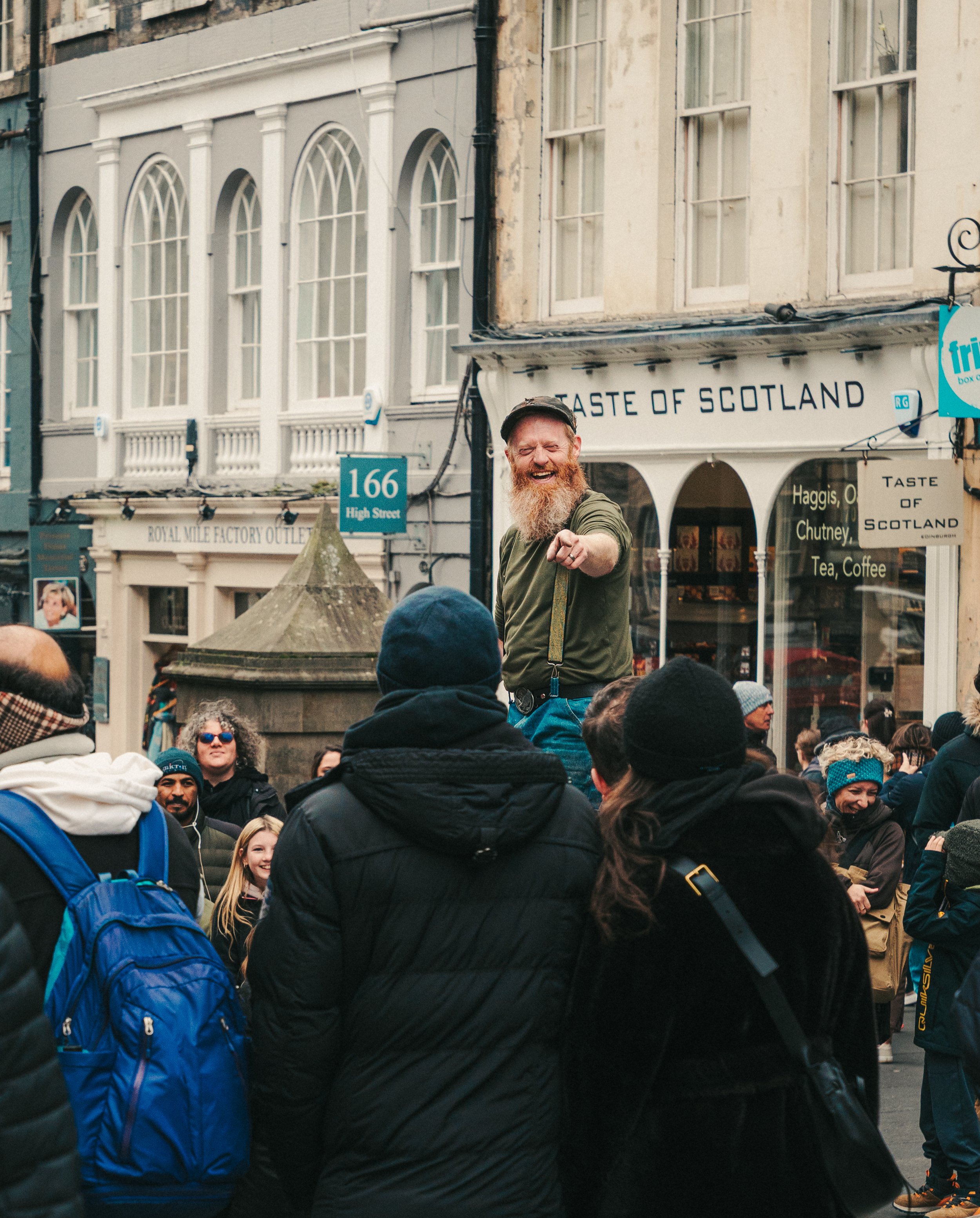 A crowd of people gathered outdoors in front of a building with large arched windows. A man with a beard and wearing a hat and green jacket is smiling and pointing towards the camera. A sign reads 'Taste of Scotland' and there are other signs and sto
