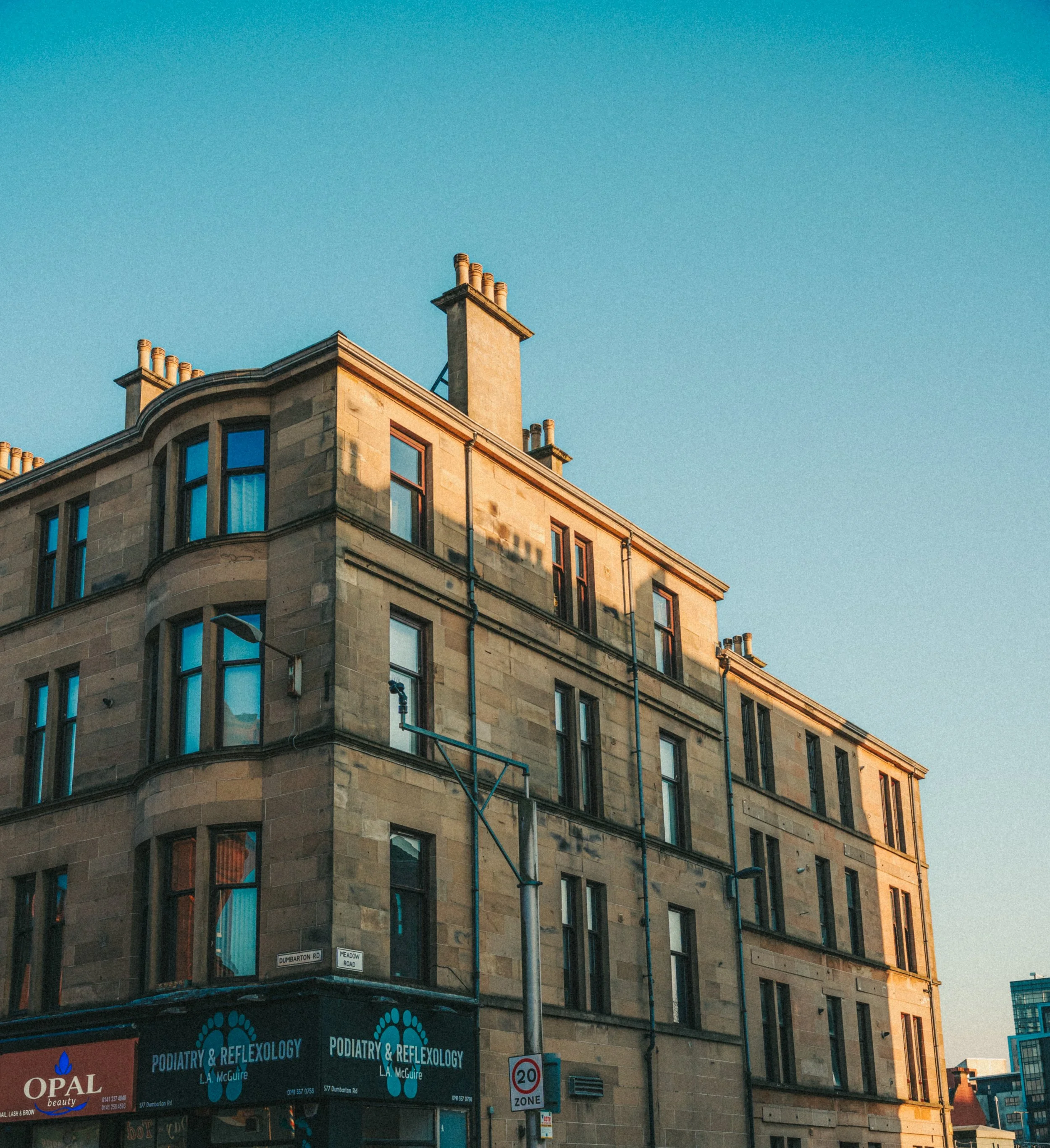 Corner of a multi-story building with stone facade, large windows, and a sign for a podiatry and reflexology clinic. There are street signs indicating Dumarton Road and Meadow Road, and a speed zone sign with a limit of 20 mph. The sky is clear and b