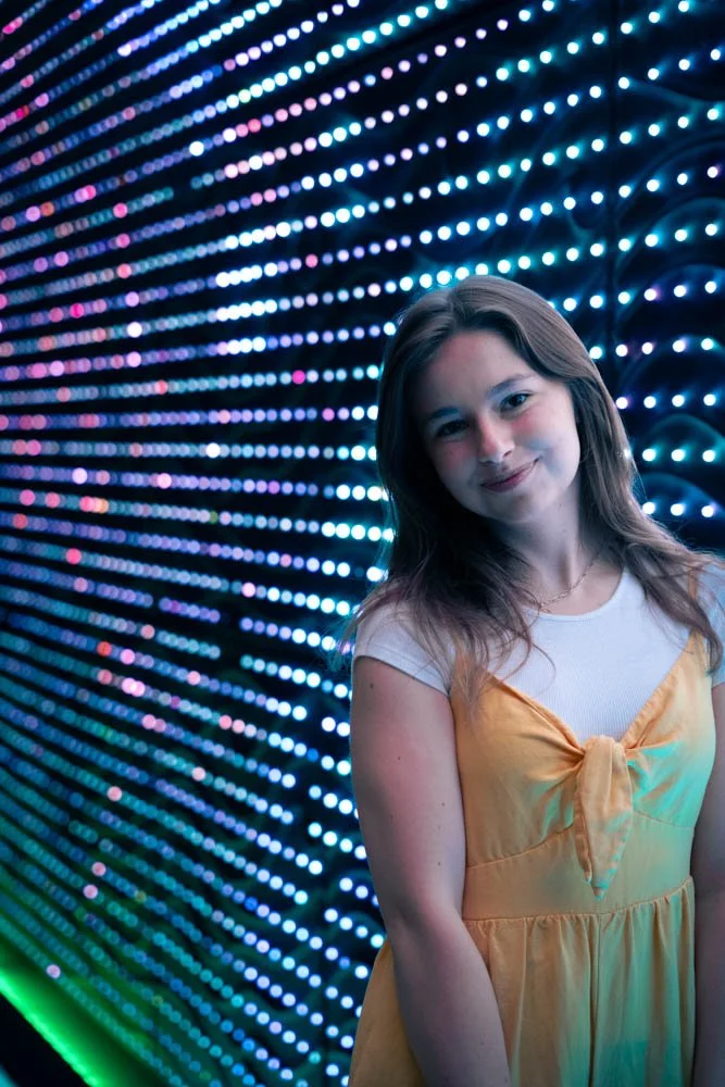 Young woman with light brown hair smiling, standing in front of a wall of colorful LED lights.