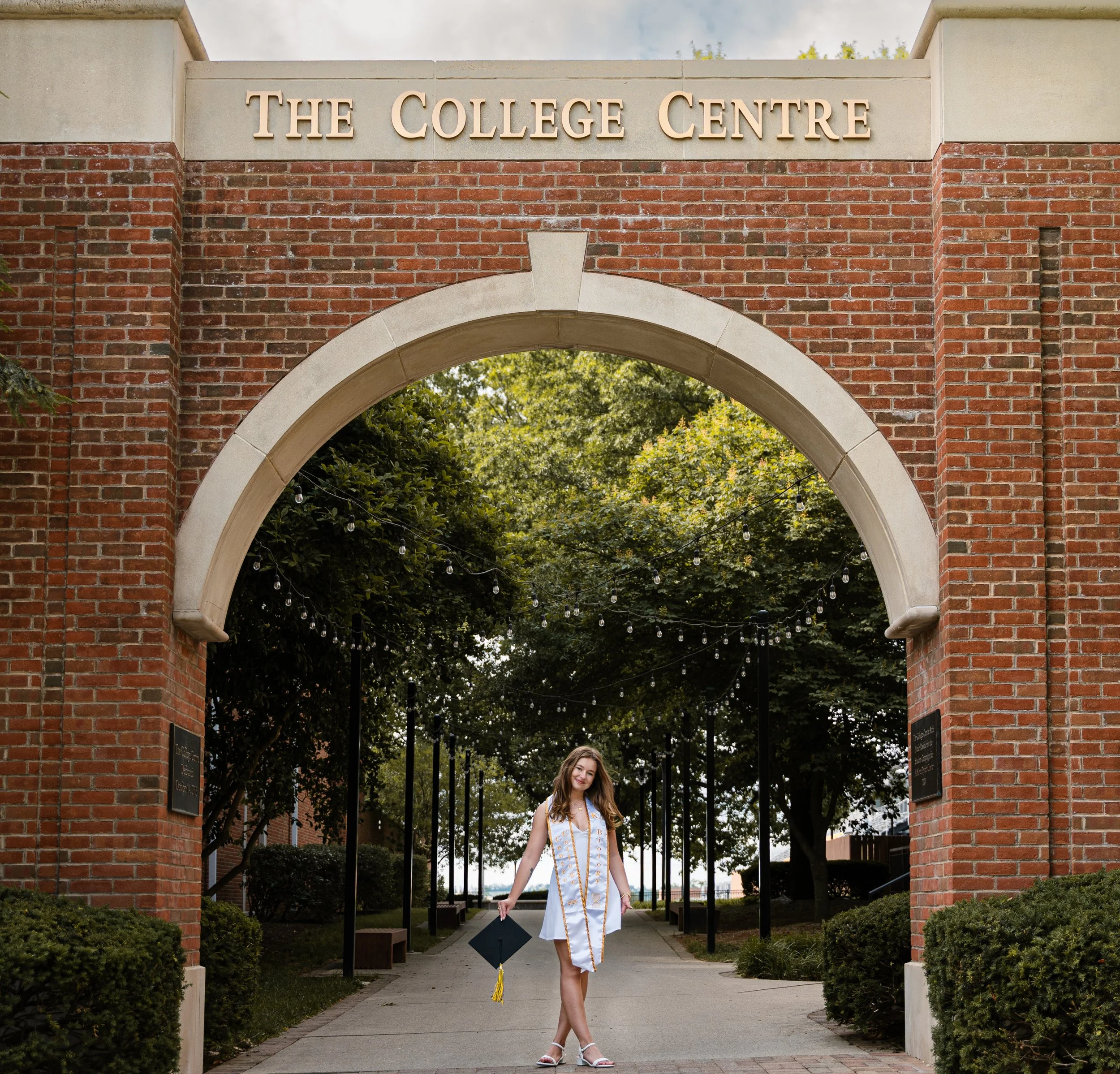 A young woman in graduation cap and gown standing under an arched brick entrance at The College Centre, holding a diploma, with trees and a cloudy sky in the background.