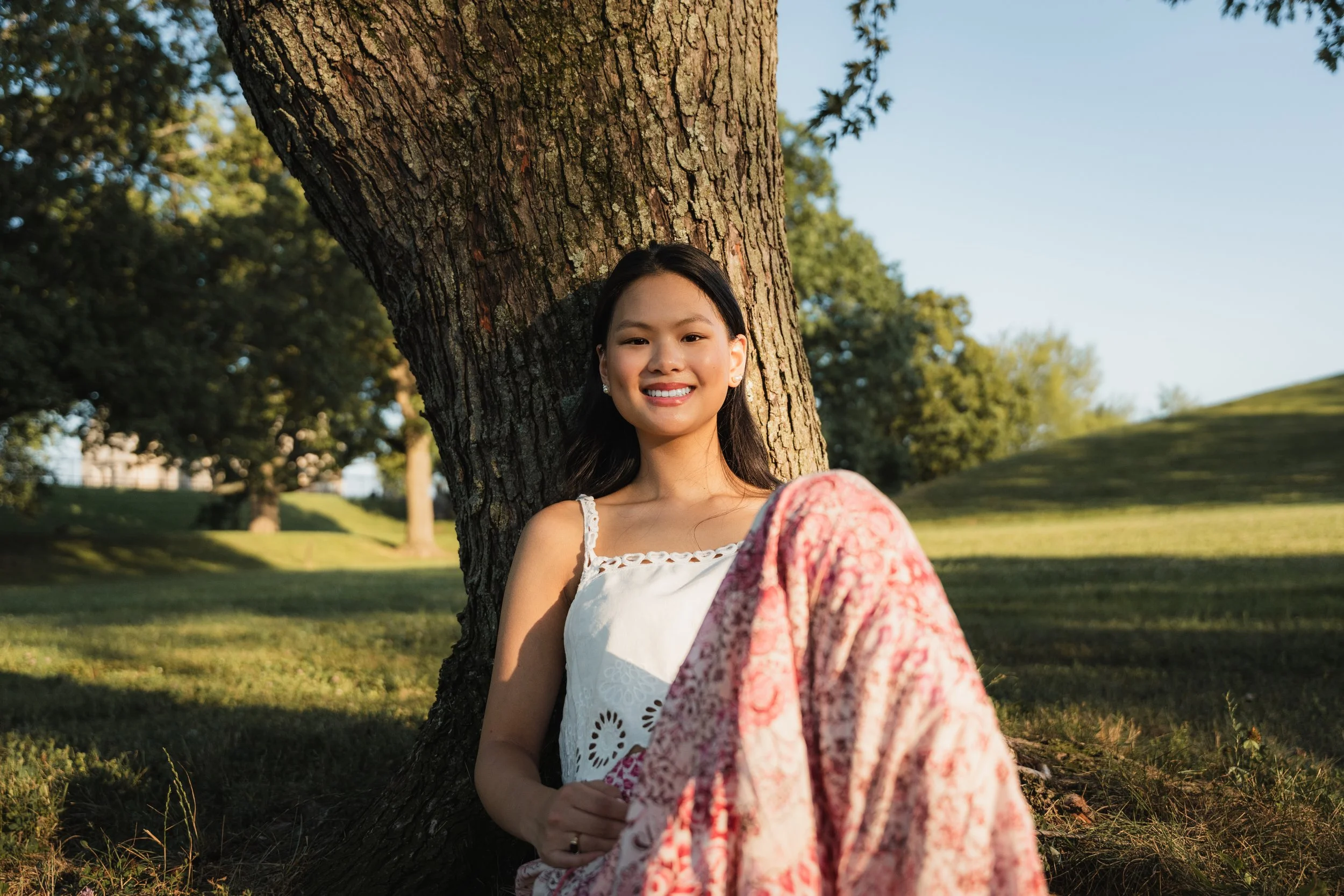 A young woman with black hair smiling and sitting by a tree in a park on a sunny day, with green grass and trees in the background.