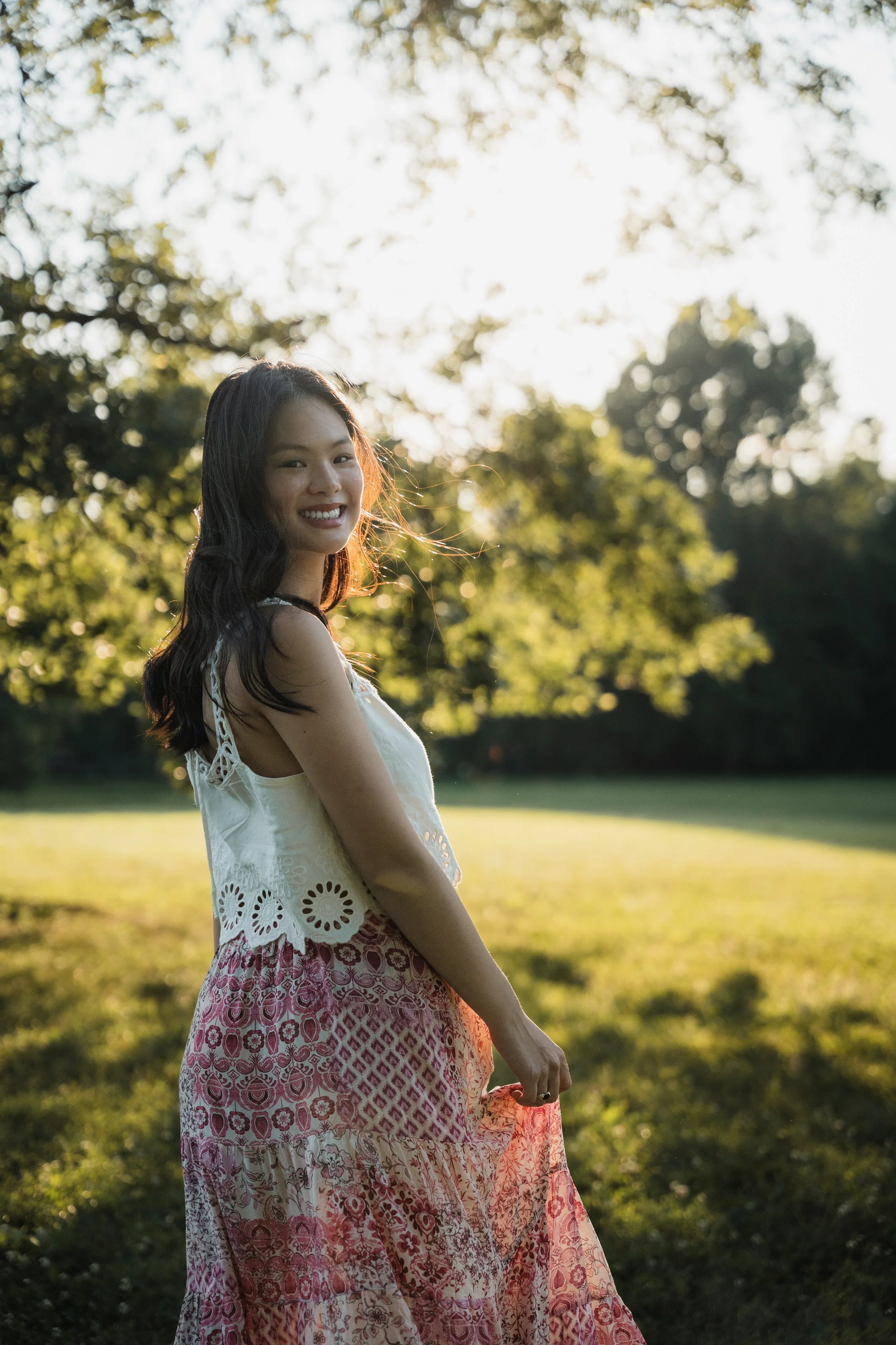 A young woman standing outdoors in a park during sunset, smiling at the camera, wearing a white sleeveless top and a patterned skirt with trees and sunlight in the background.