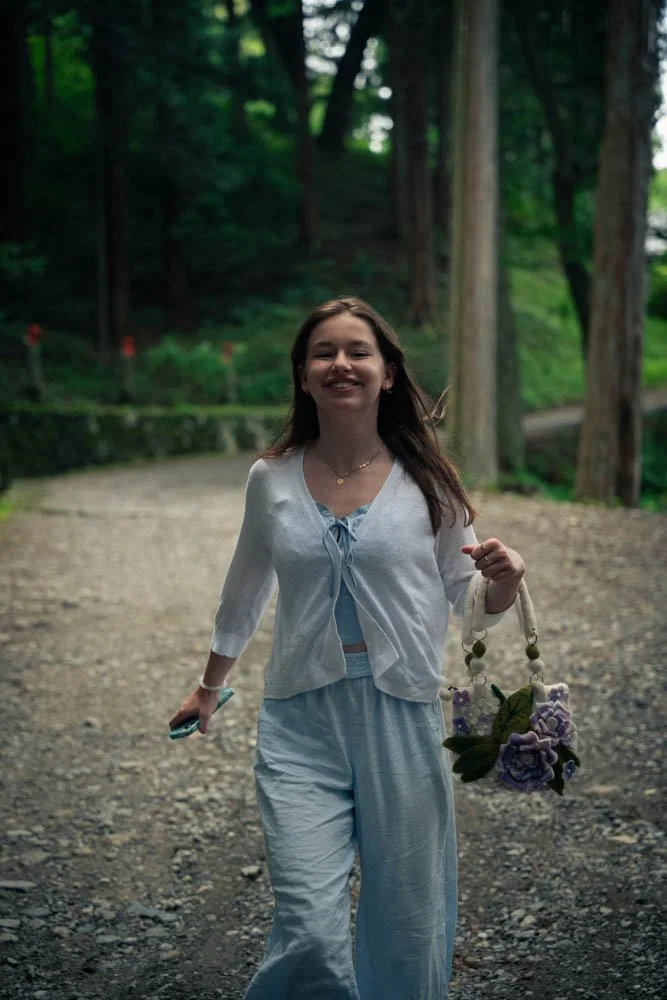 A young woman smiling while walking outdoors on a gravel path in a forest, carrying a small floral bag.