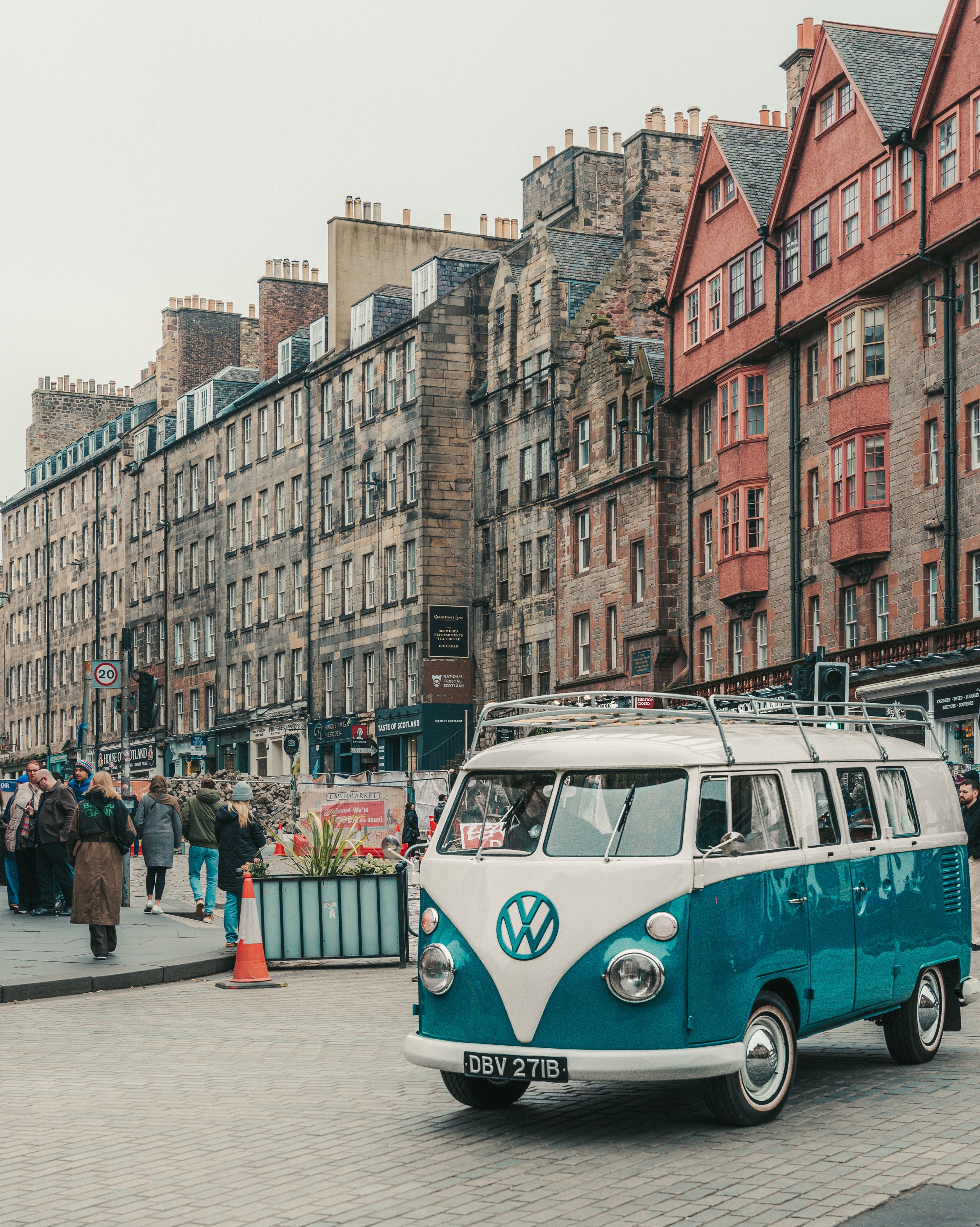A crowded street scene with a vintage blue and white Volkswagen van in the foreground, people walking and talking, old stone buildings with multiple chimneys in the background, and various signs and storefronts along the street.
