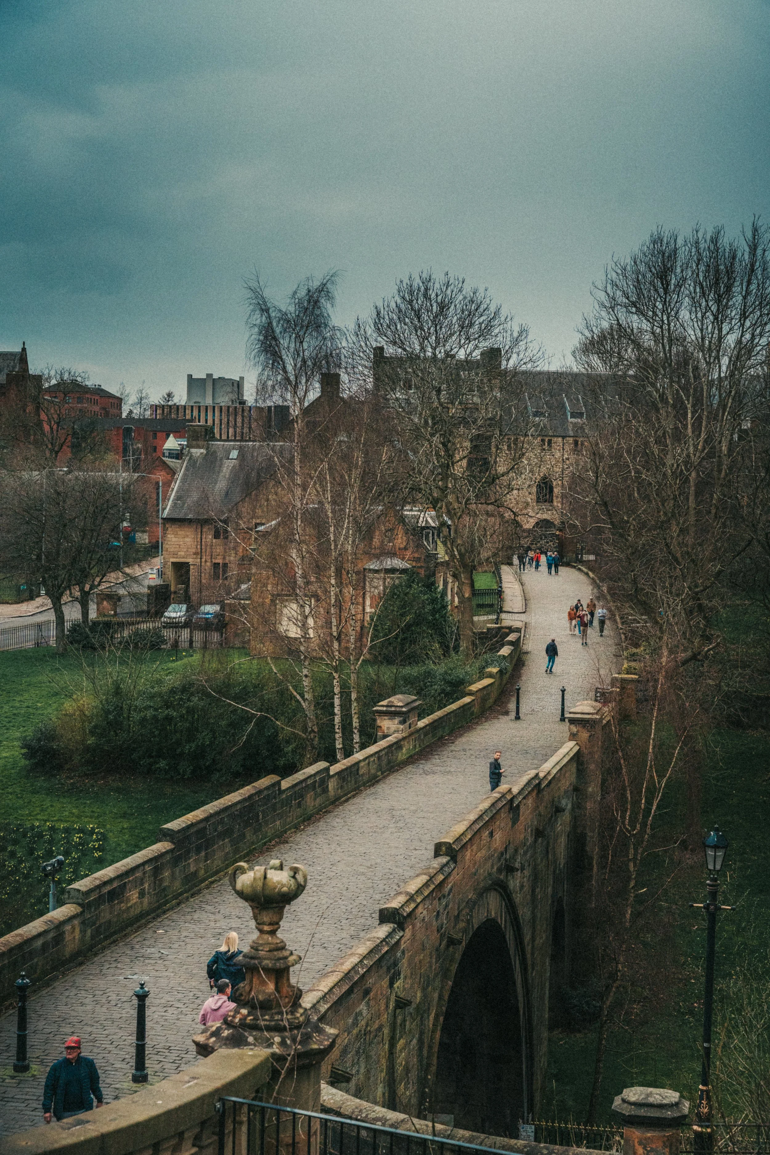 A stone bridge with a fountain and several people walking across it, leading to a path with trees and historic buildings in the background.