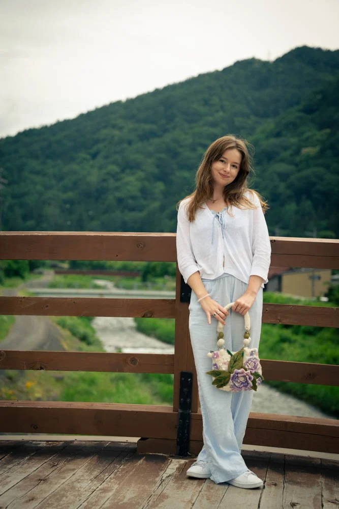 Young woman standing on a wooden bridge with a green mountain and river in the background, holding a flower-shaped bag, wearing a white top and light blue pants.