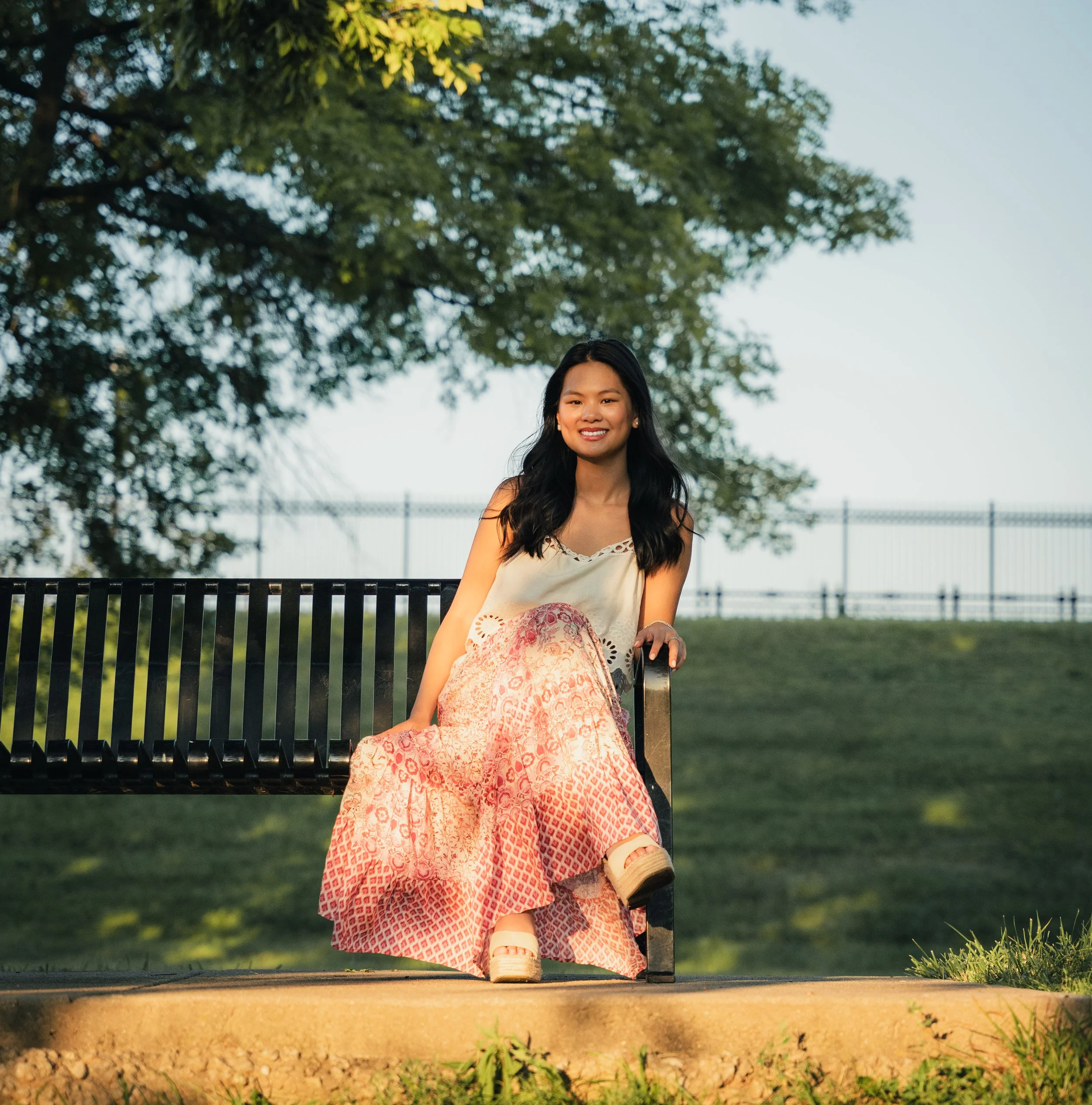 A young woman with long dark hair sitting on a park bench smiling, wearing a white sleeveless top and a pink patterned skirt, with a large green tree behind her and a fence in the distance.