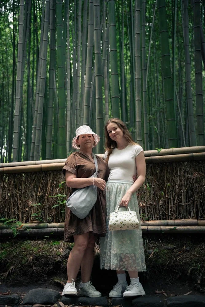 Two women standing in front of a bamboo forest, one wearing a brown dress and white hat, the other in a white top and patterned skirt, both holding bags.