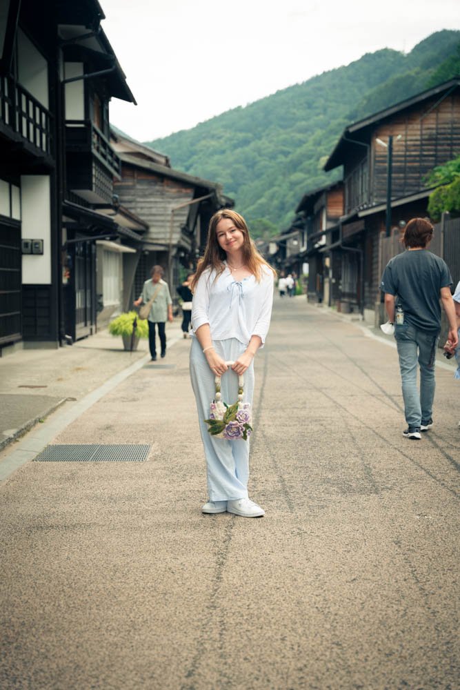 A young woman stands in the middle of a street lined with traditional Japanese wooden buildings, holding a floral handbag, with a mountain in the background.