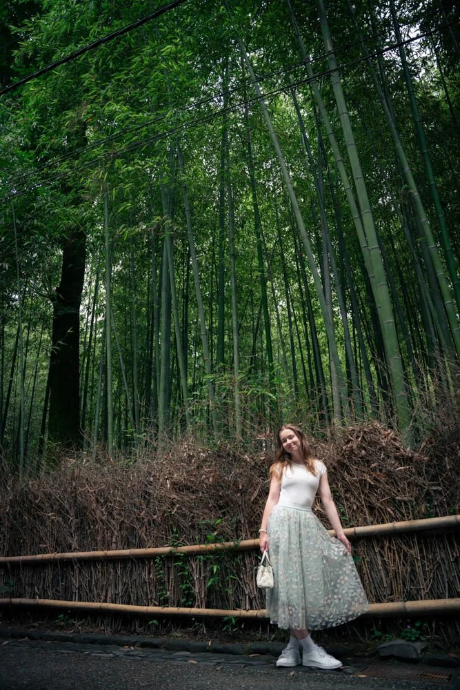 A young woman in a white sleeveless top and a long, floral, semi-transparent skirt standing on a sidewalk in front of a bamboo forest, holding a small purse and smiling.