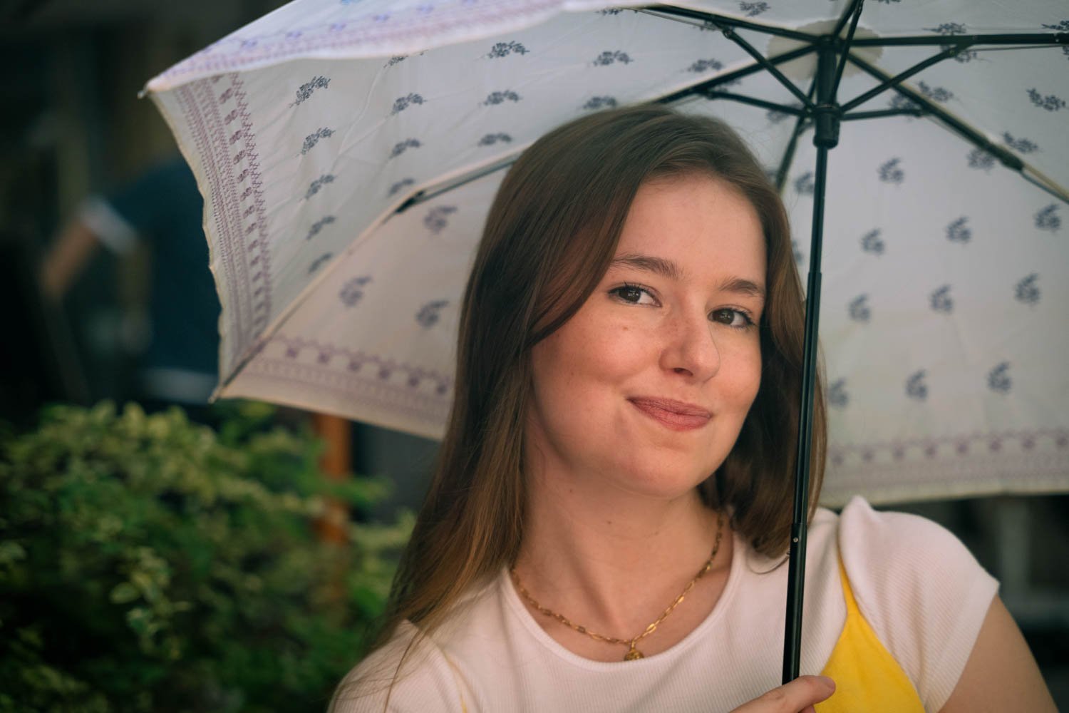 A young woman with brown hair and a yellow and white shirt holding a patterned umbrella outdoors.