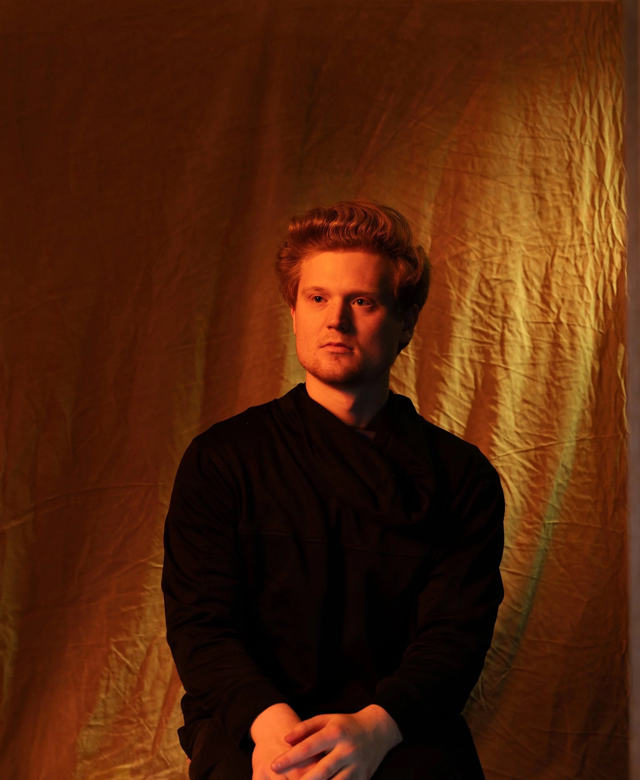 A young man with reddish-brown hair and light facial hair, wearing a black shirt, sitting with arms crossed against a textured gold curtain backdrop, lit warmly from the side.