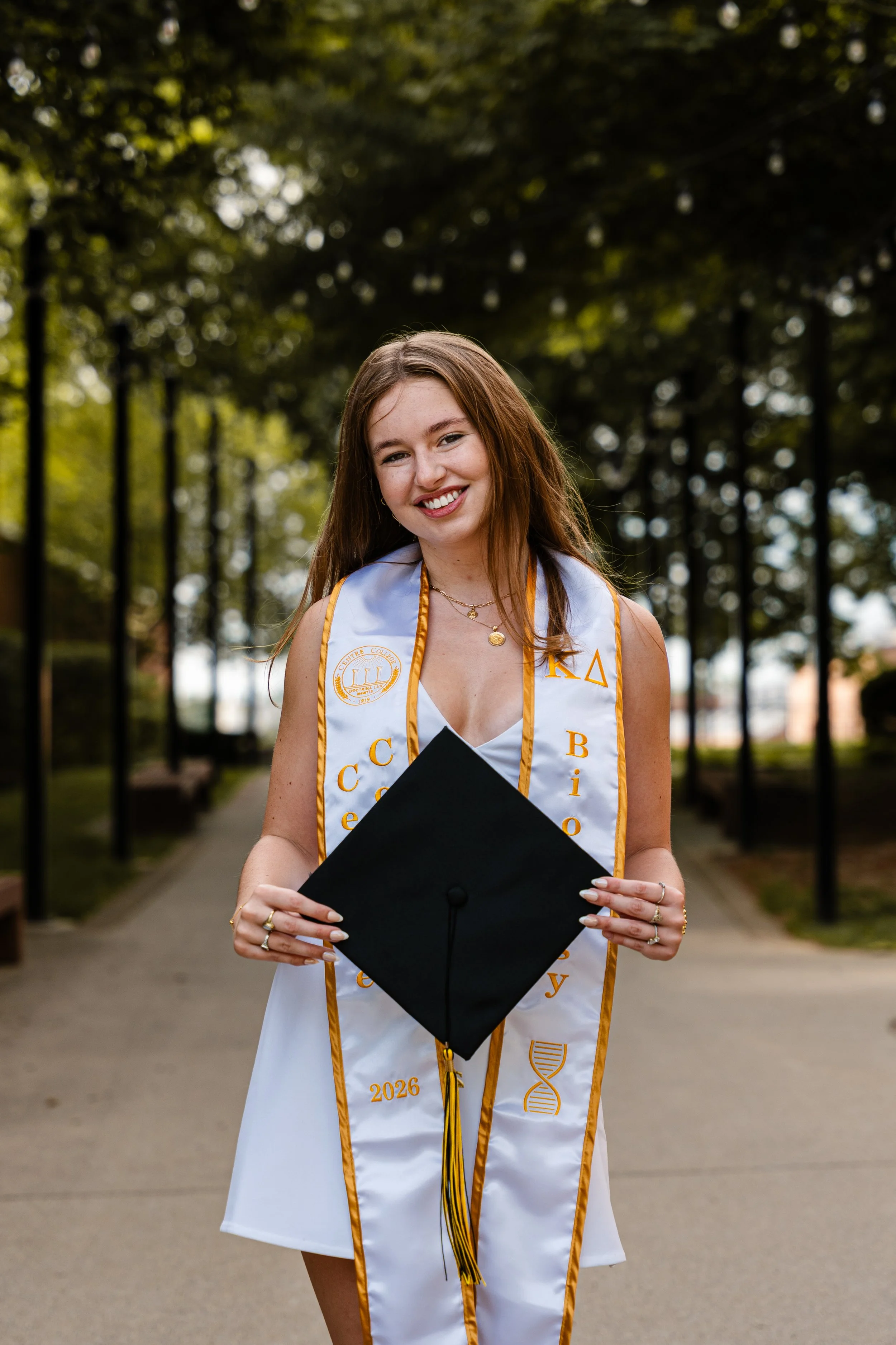 A young woman in a white graduation dress and sash holding a black graduation cap, standing outdoors on a pathway with trees in the background.