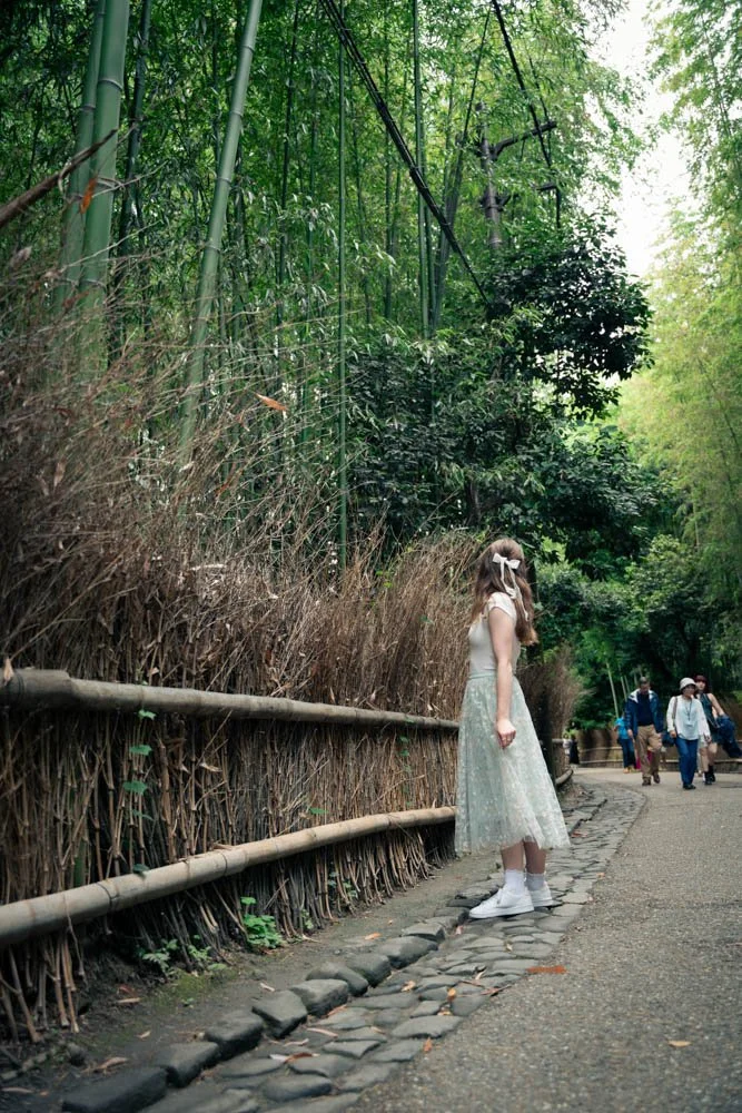A girl with long hair, wearing a white dress and white sneakers, stands on a pebble path next to a bamboo fence in a lush green forest, with a few people walking further down the path.